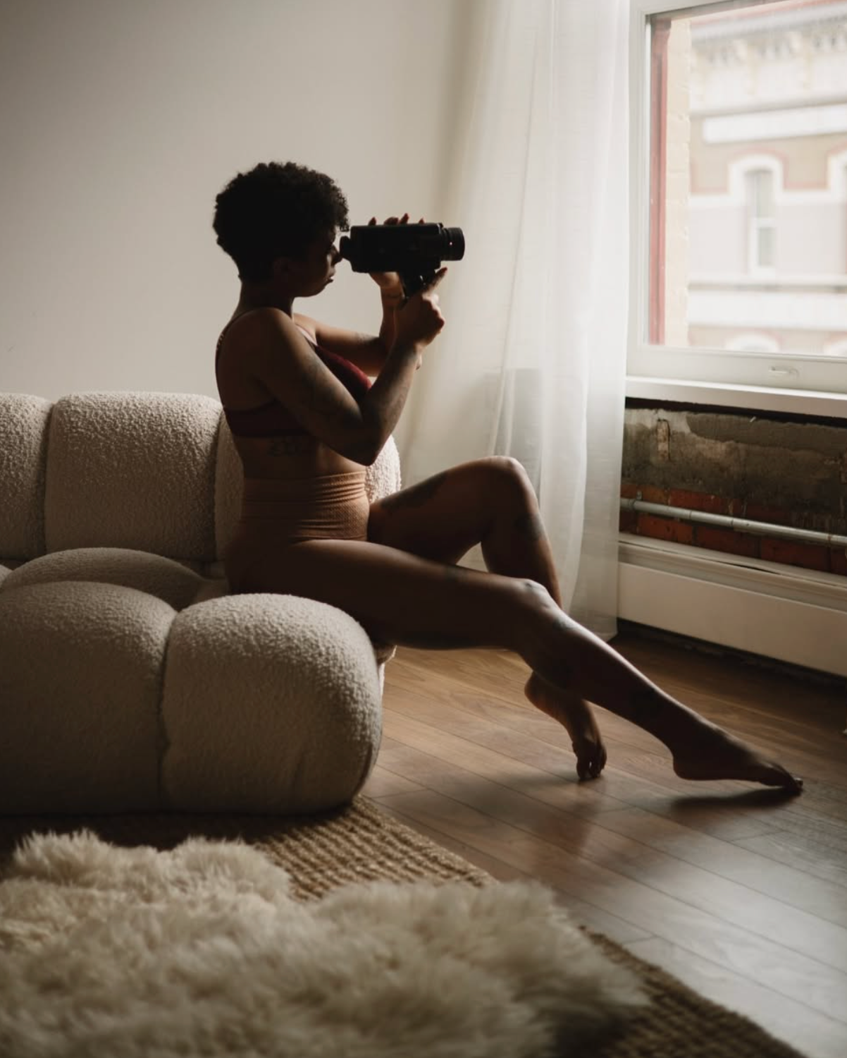 Silhouetted figure with camera sitting on plush couch near window in natural light.