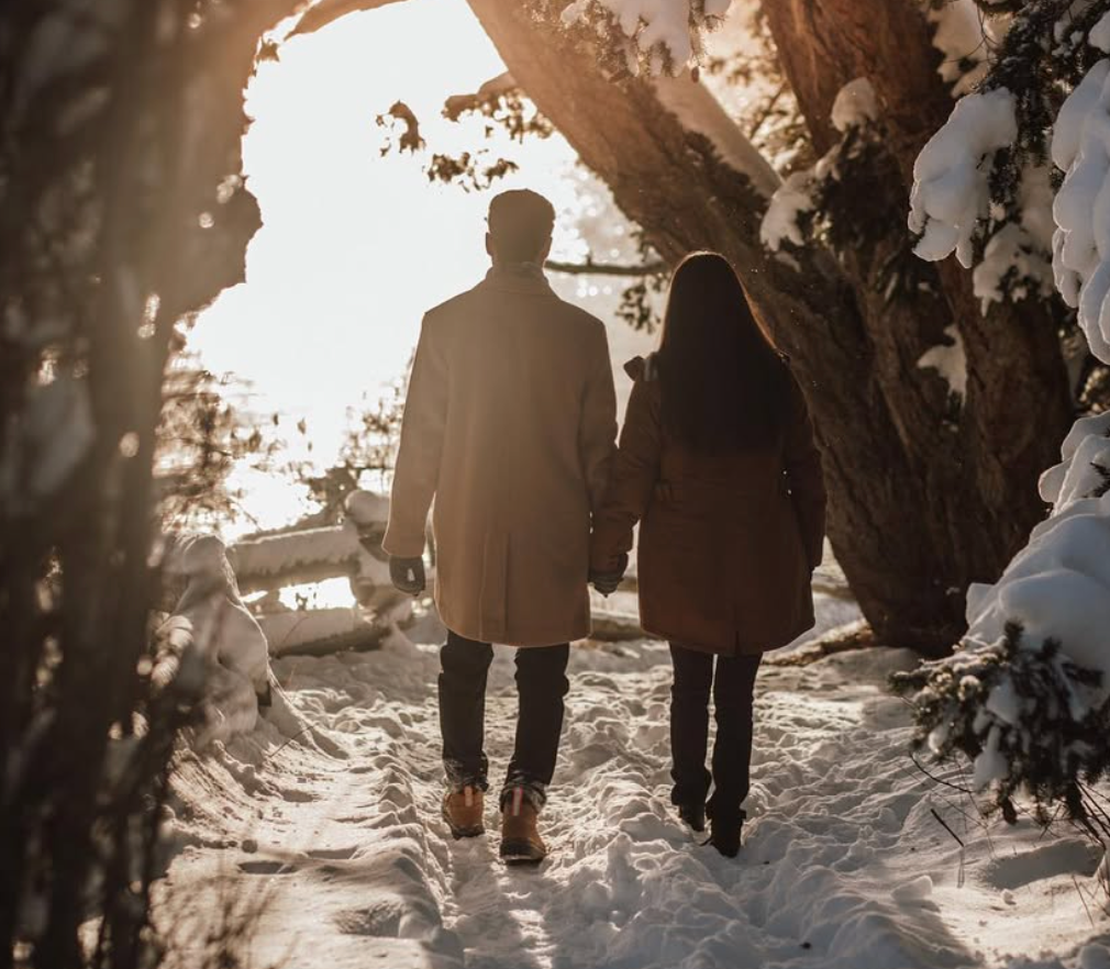 A silhouetted couple walks hand in hand through a snowy winter landscape at sunset.