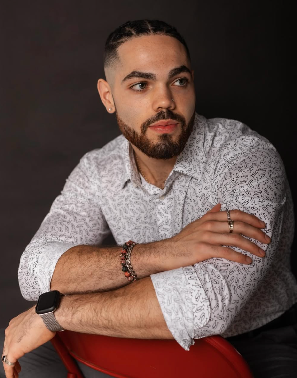 Man in patterned shirt poses confidently against dark backdrop wearing jewelry.