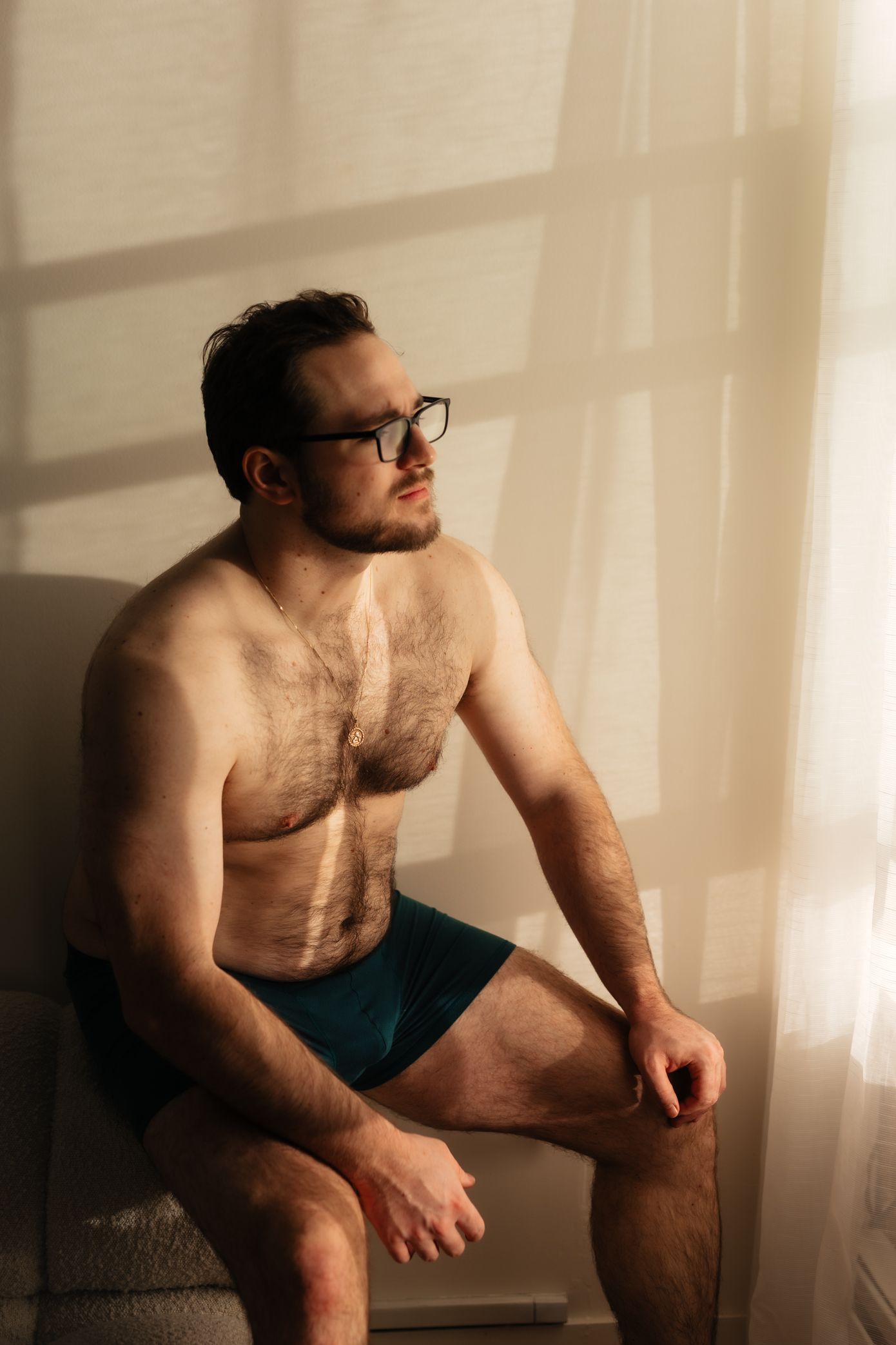 Shirtless man in glasses and teal underwear sitting by a sunlit curtain window in a moody indoor setting.