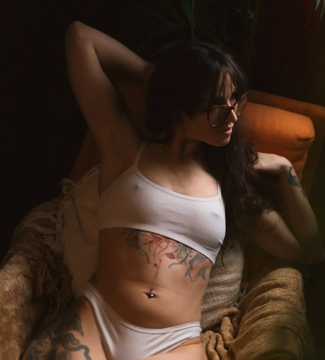 Artistic boudoir photo showing torso with floral tattoo in white crop top and cardigan in moody lighting.