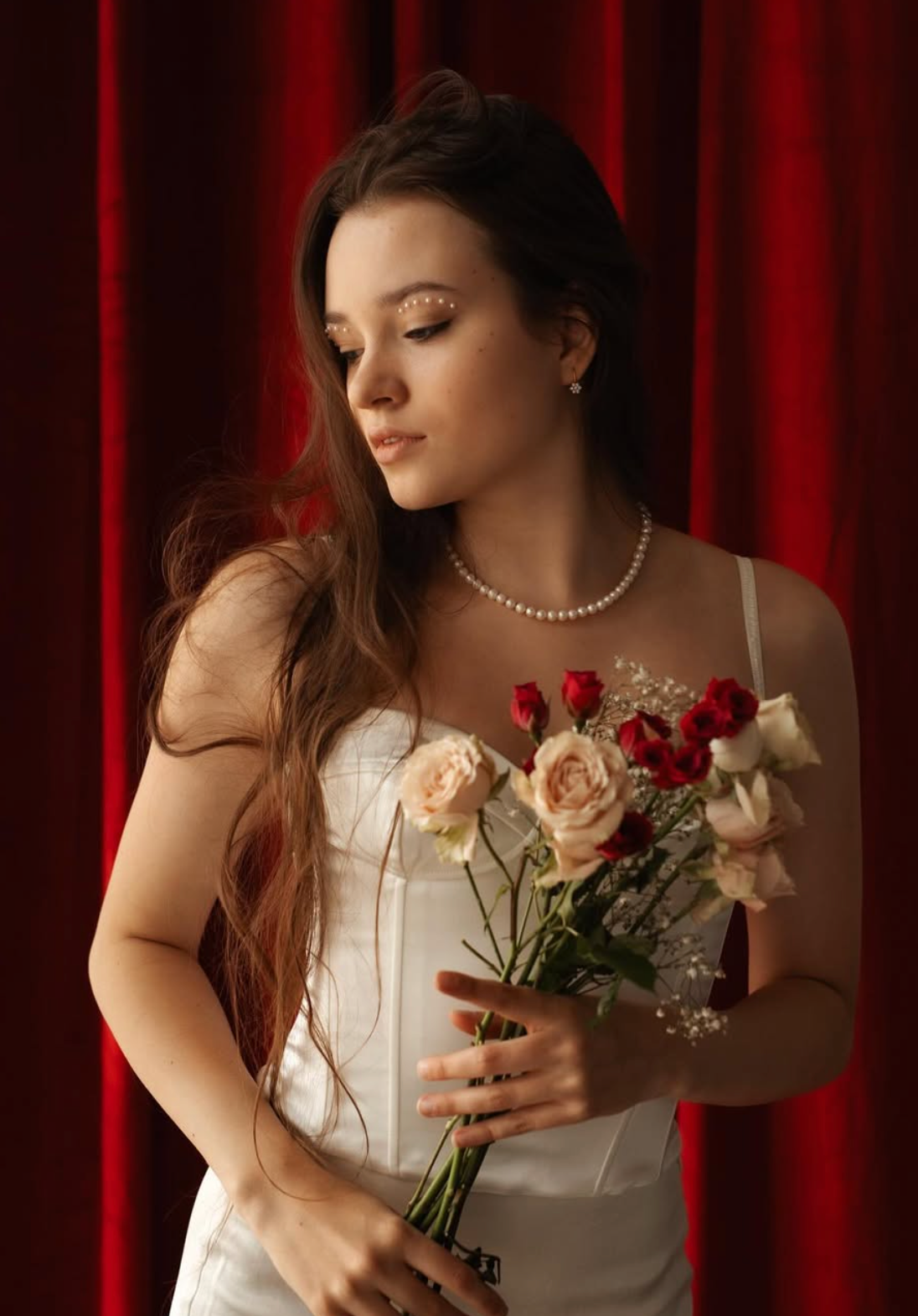 Woman in white dress holding roses against red curtain backdrop in dramatic lighting.
