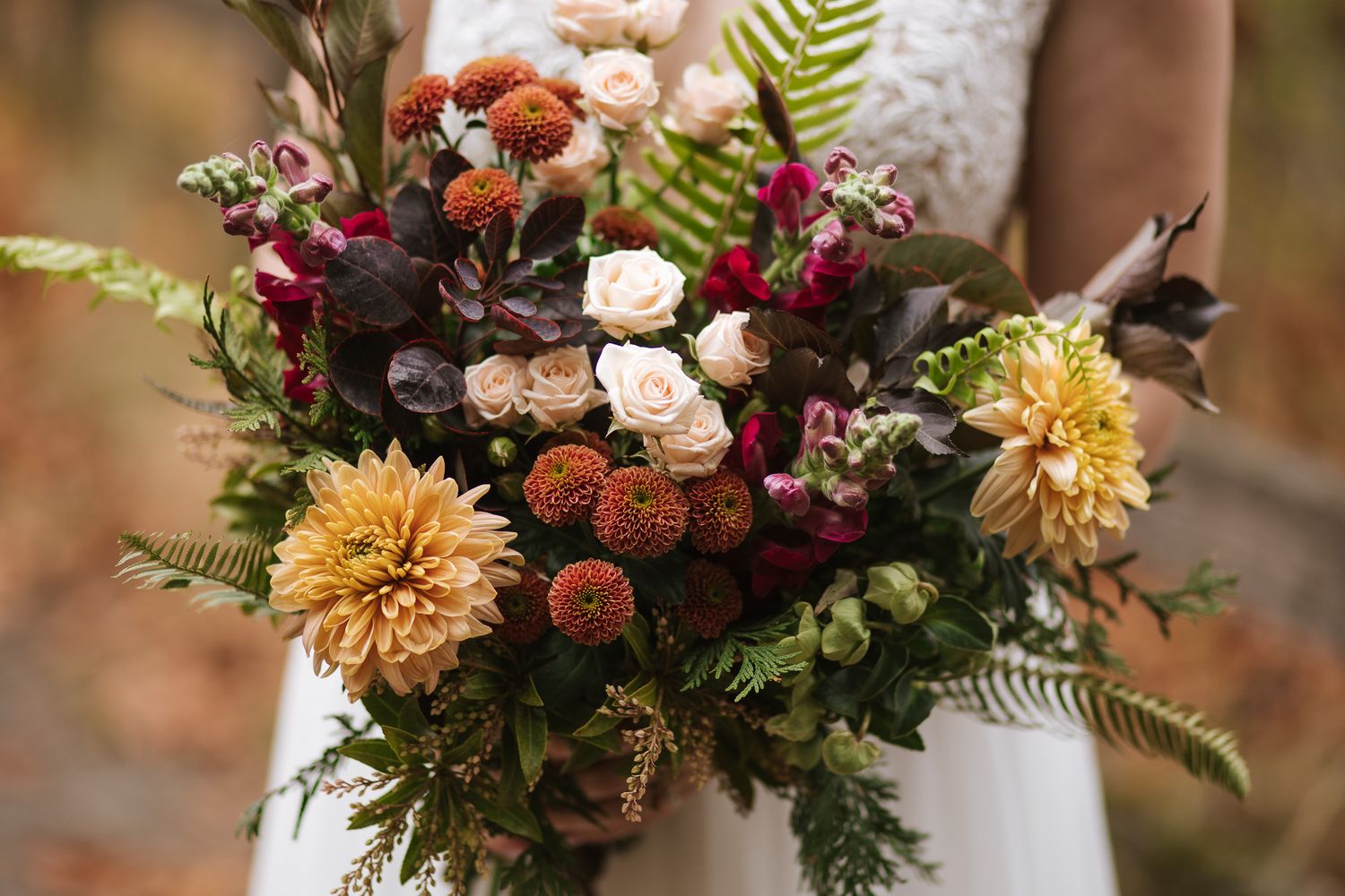 Rustic autumn wedding bouquet with orange dahlias, burgundy flowers, and green ferns arranged in natural style.