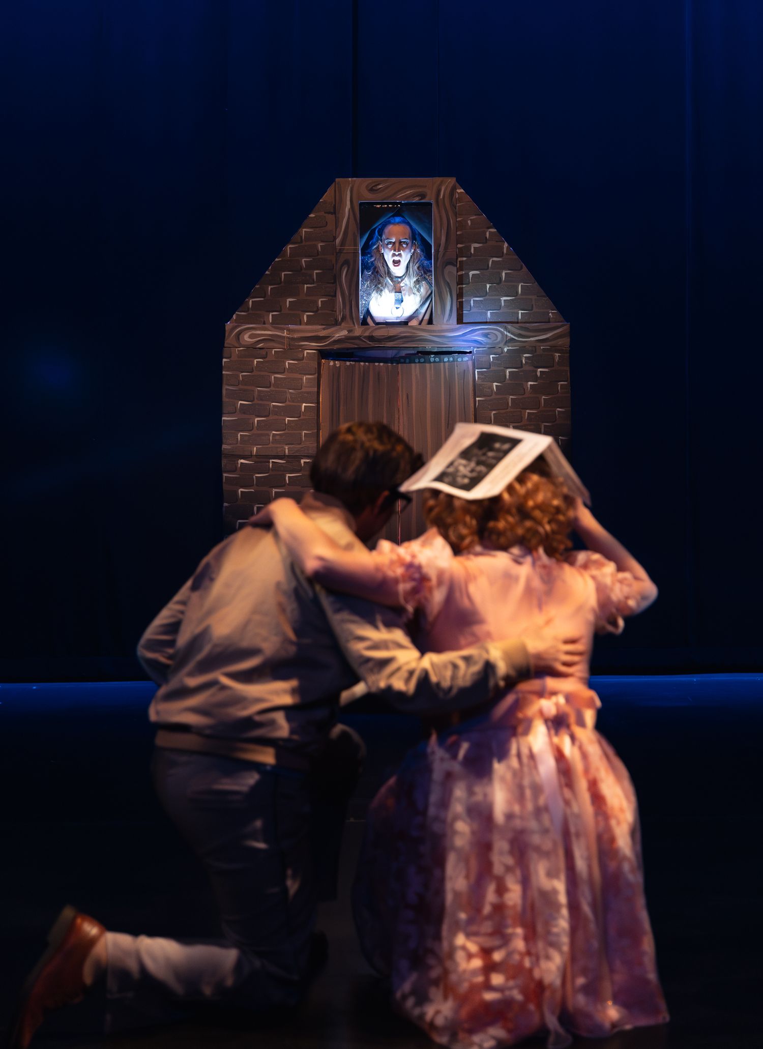Stage actors embrace while looking at illuminated portrait in brick alcove during theatrical performance.