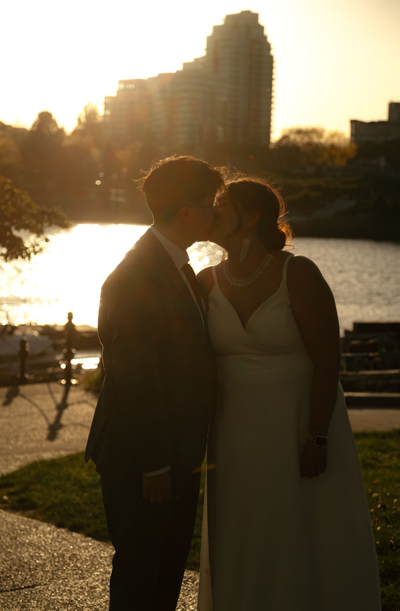Silhouetted couple embraces during golden hour by the waterfront as sun sets behind city skyline.