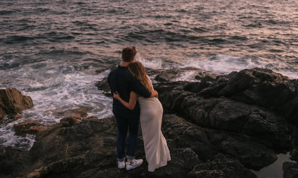 Couple embracing while watching sunset on rocky ocean shore.