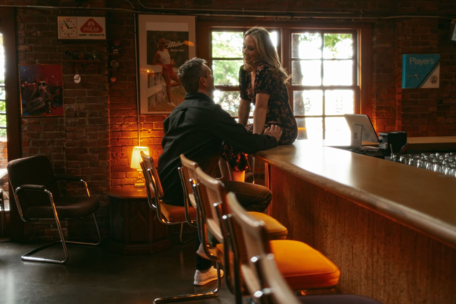 Two people conversing at a rustic brick-walled bar with warm lighting and vintage decor.