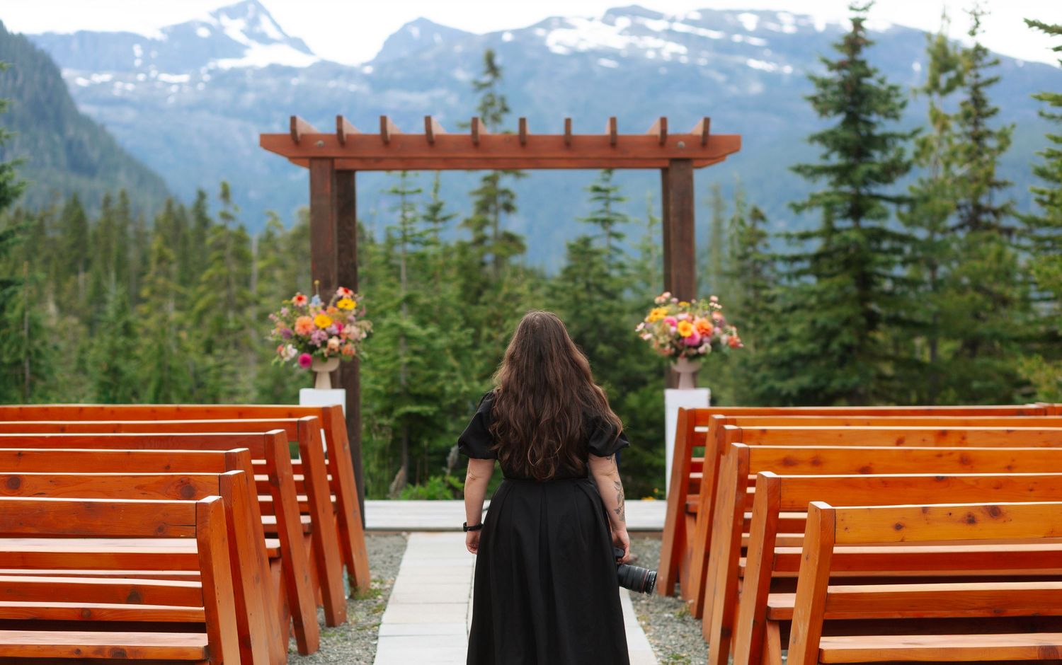Outdoor wedding ceremony setup with wooden pews, floral arrangements, pergola altar, and scenic mountain forest backdrop.