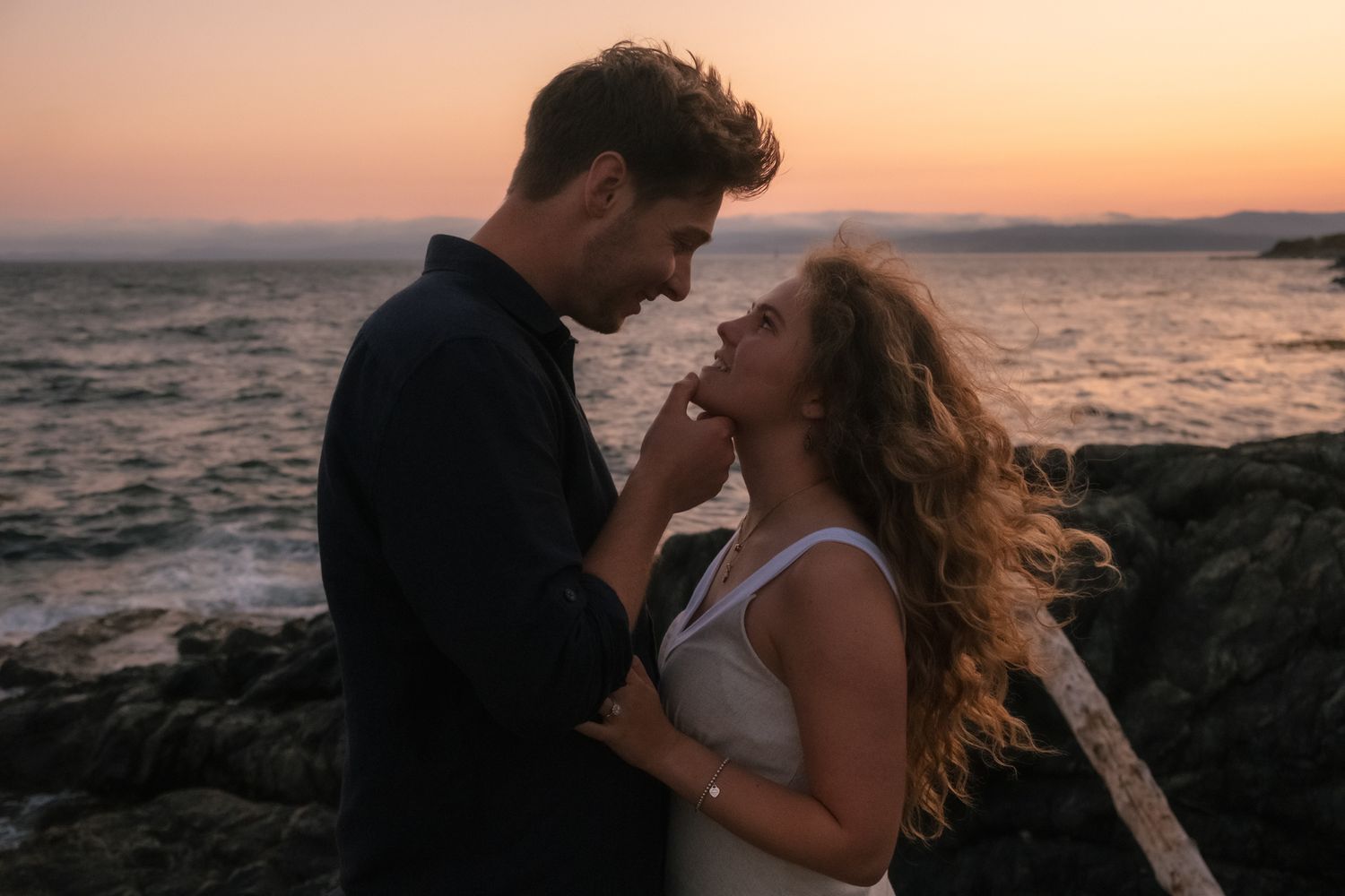 Couple sharing a romantic moment at sunset by rocky ocean shore, warm pink sky in background.