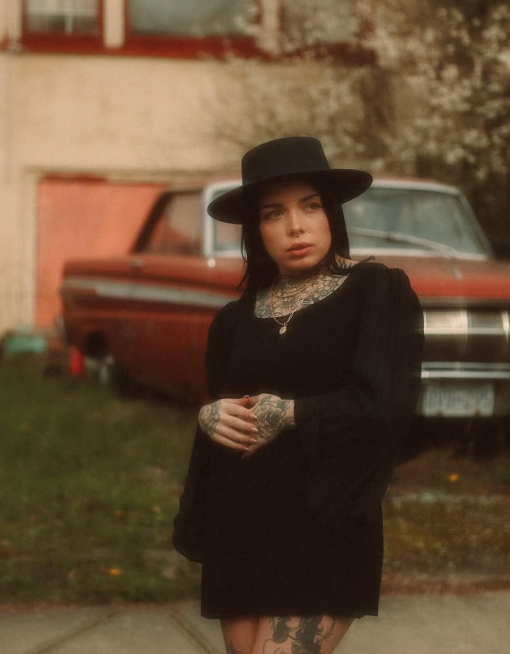 Vintage-styled portrait against classic car featuring black dress and wide-brimmed hat.