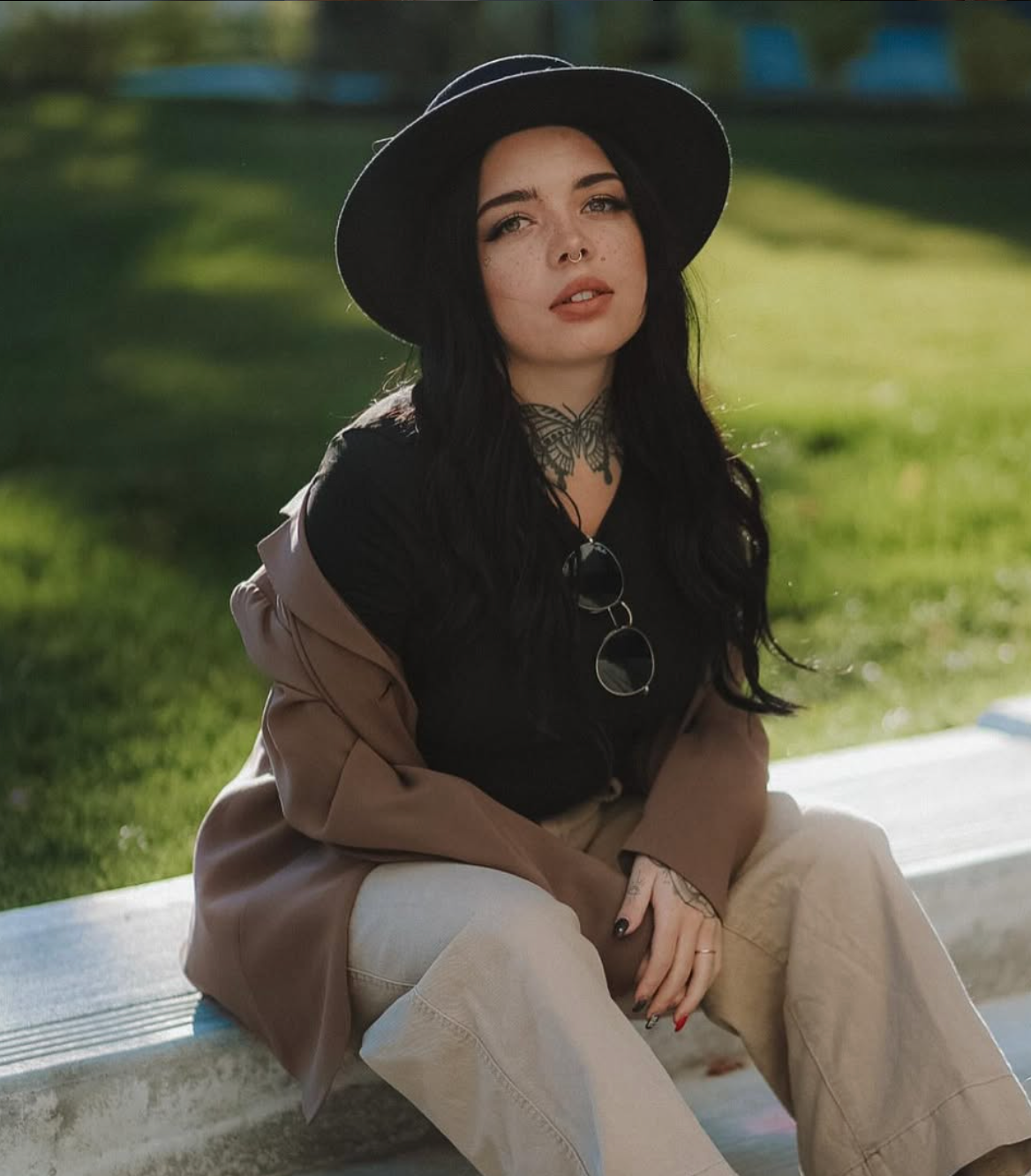 Stylish figure in brown coat and black hat sits outdoors on concrete ledge in natural sunlight.
