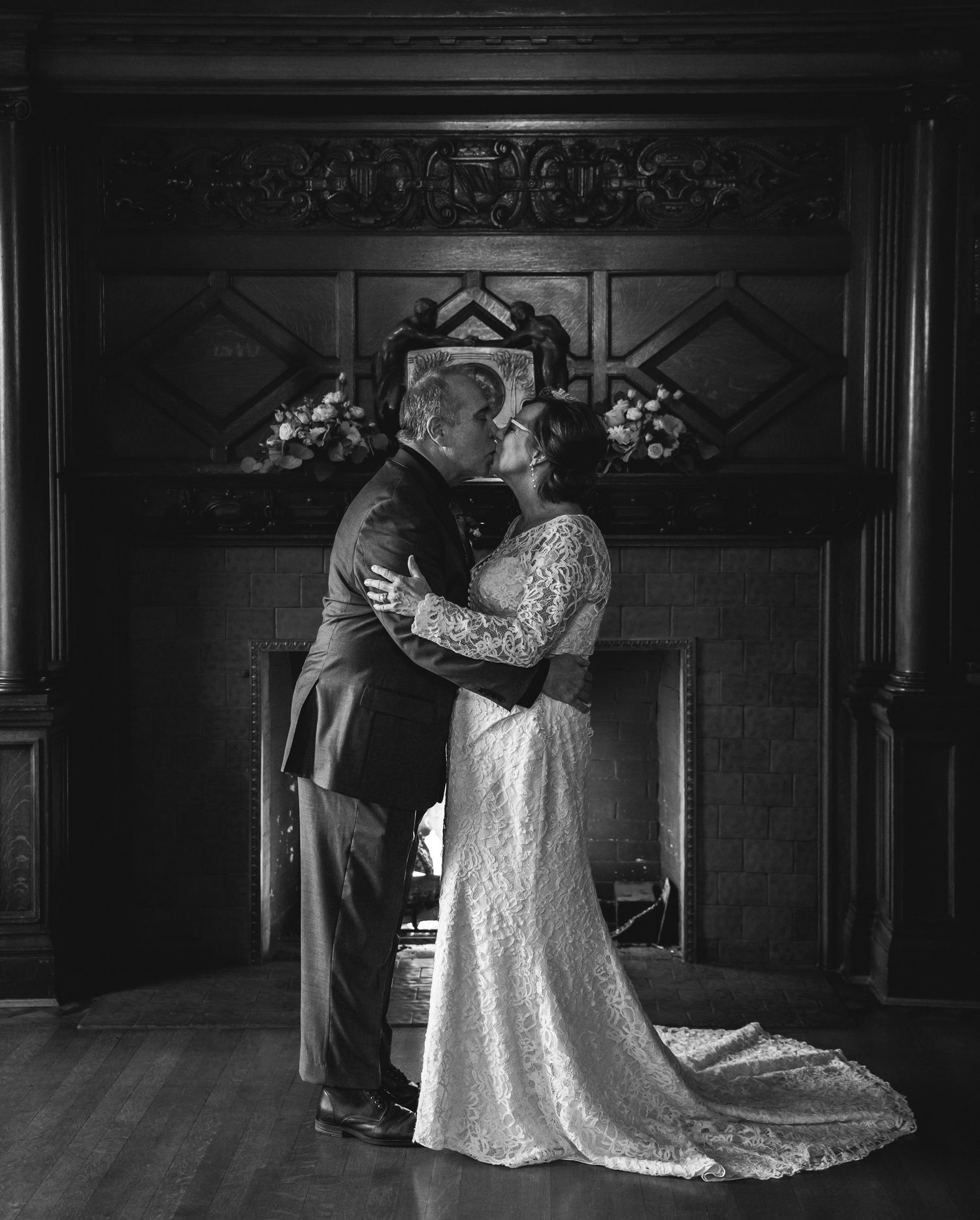 A black and white wedding photo showing a romantic moment in front of an ornate fireplace.