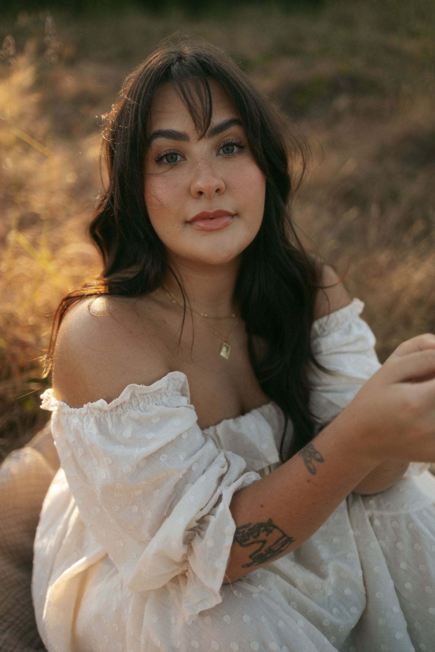 A woman in a flowing white dress poses outdoors during golden hour with warm sunlight creating a dreamy atmosphere.