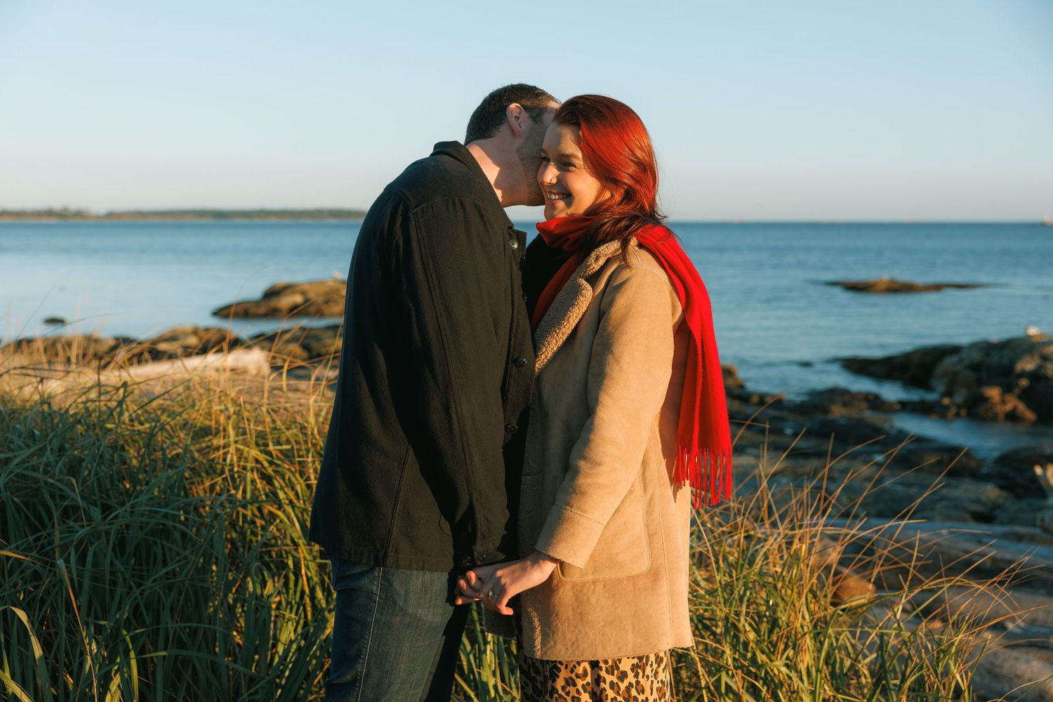 A romantic couple embraces on a coastal cliff at sunset with ocean waves crashing against rocky shores in the background.