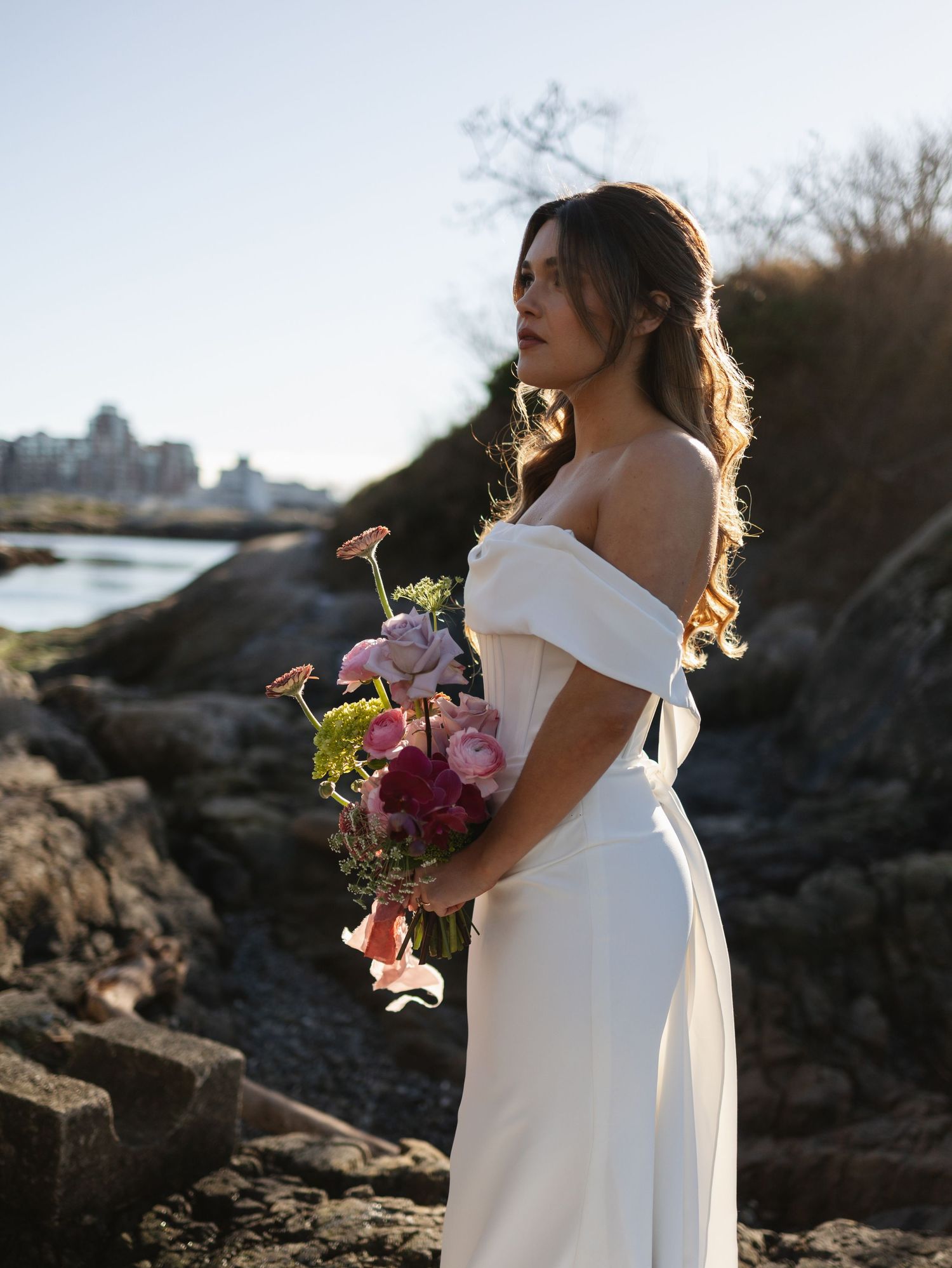 Bride in off-shoulder white gown holds colorful bouquet on rocky shore, gazing into distance with soft natural light.
