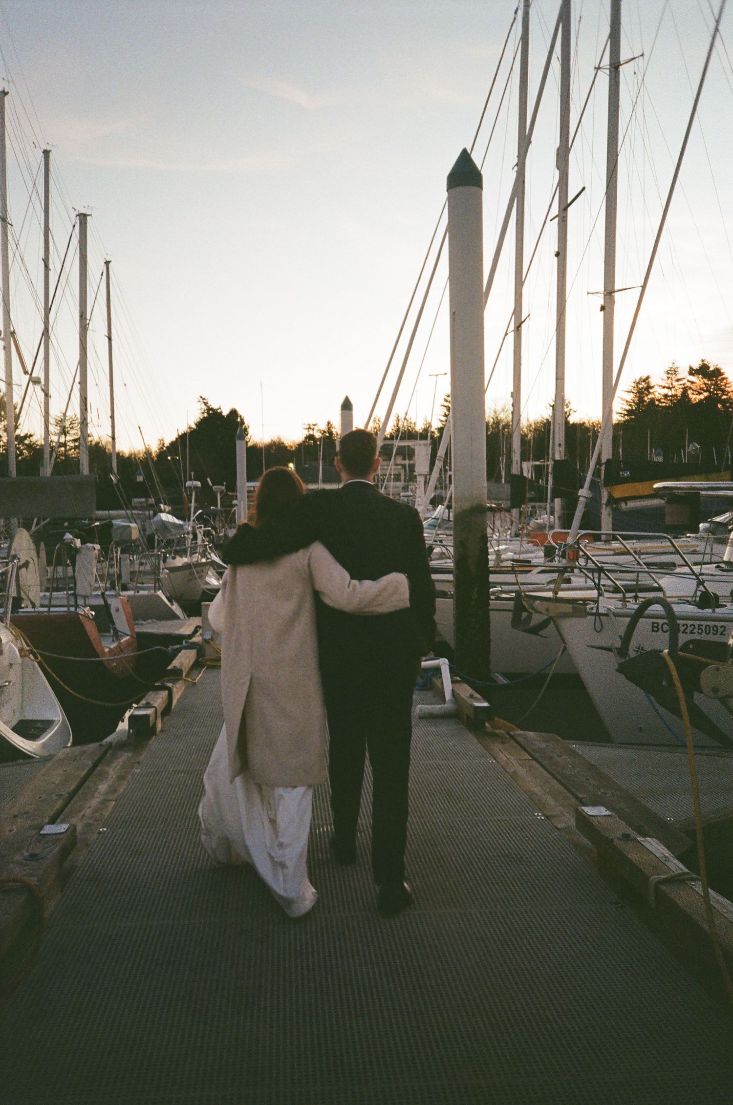A couple embraces while walking along a marina dock at sunset among sailboats and masts.