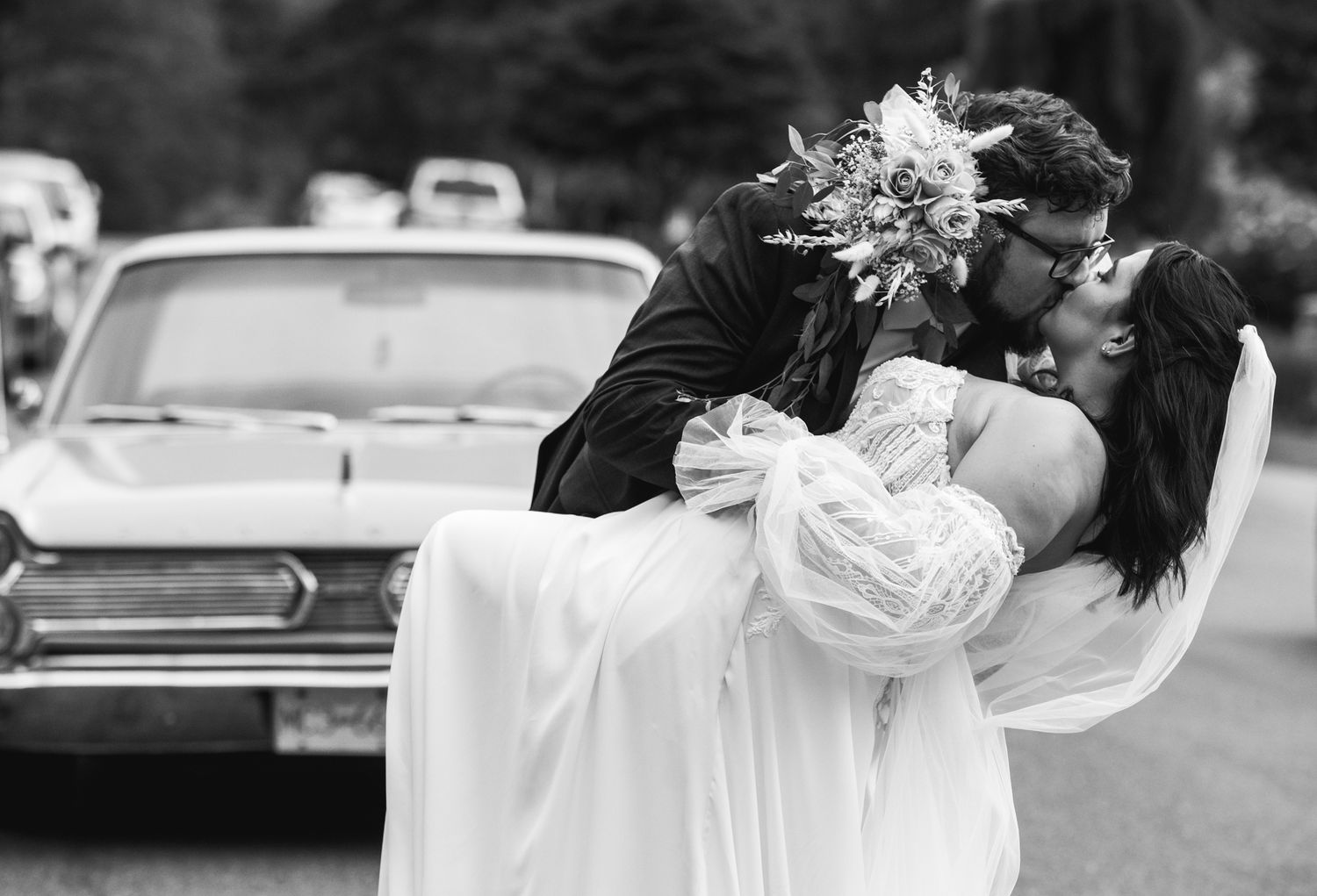 Romantic black and white wedding photo of a couple sharing an intimate moment by a vintage car.