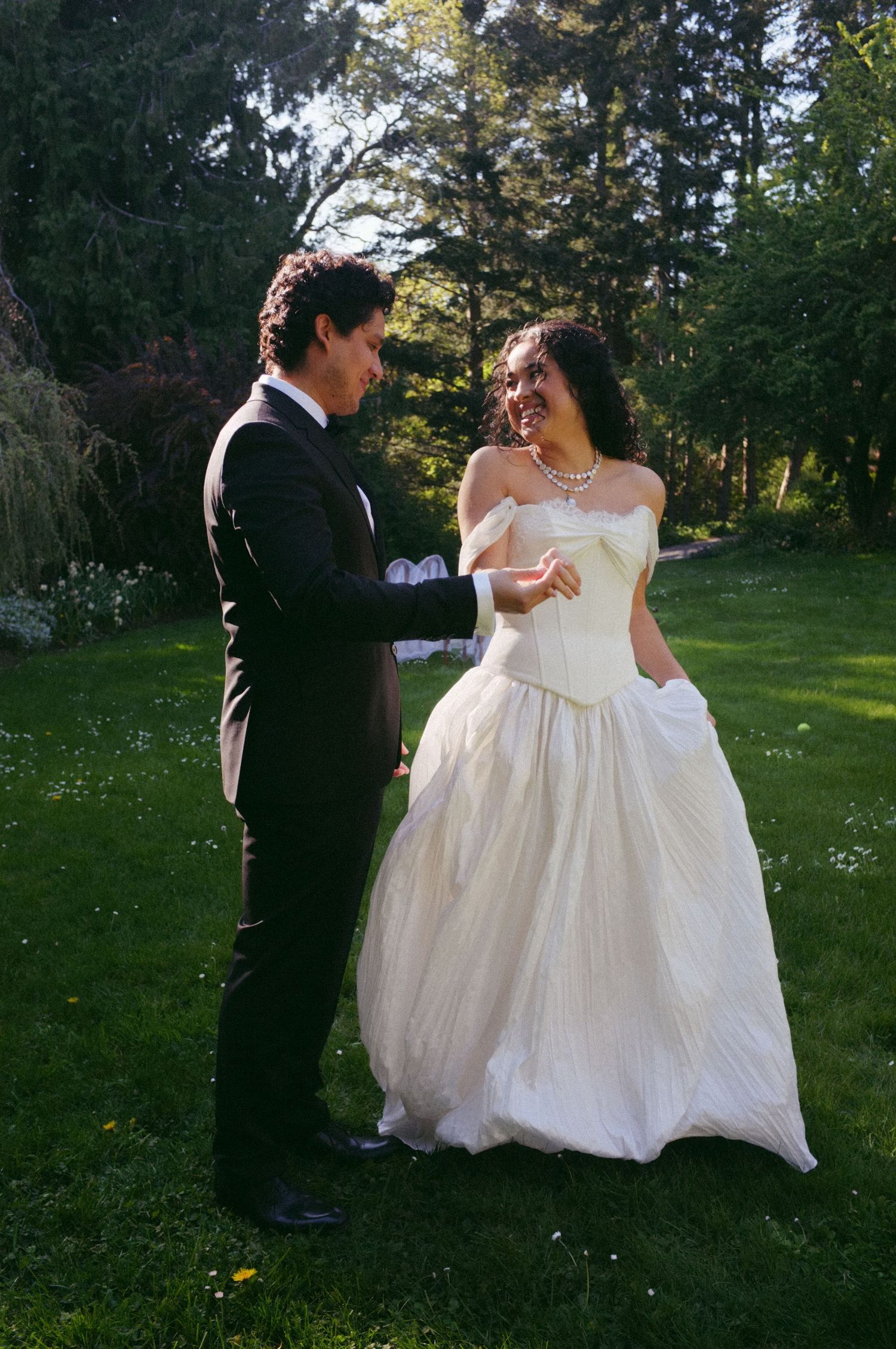 Bride in white off-shoulder ball gown and groom in black tuxedo share a joyful moment on green lawn.