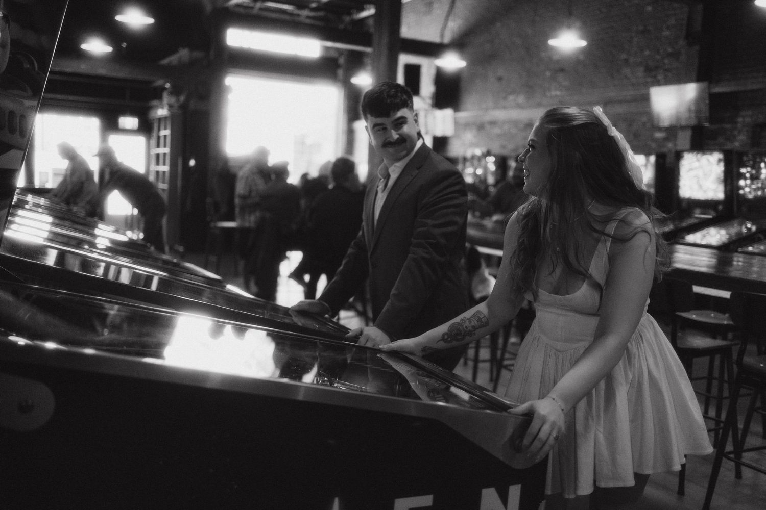 Black and white photo of a couple playing pinball together in a dimly lit bar or arcade.