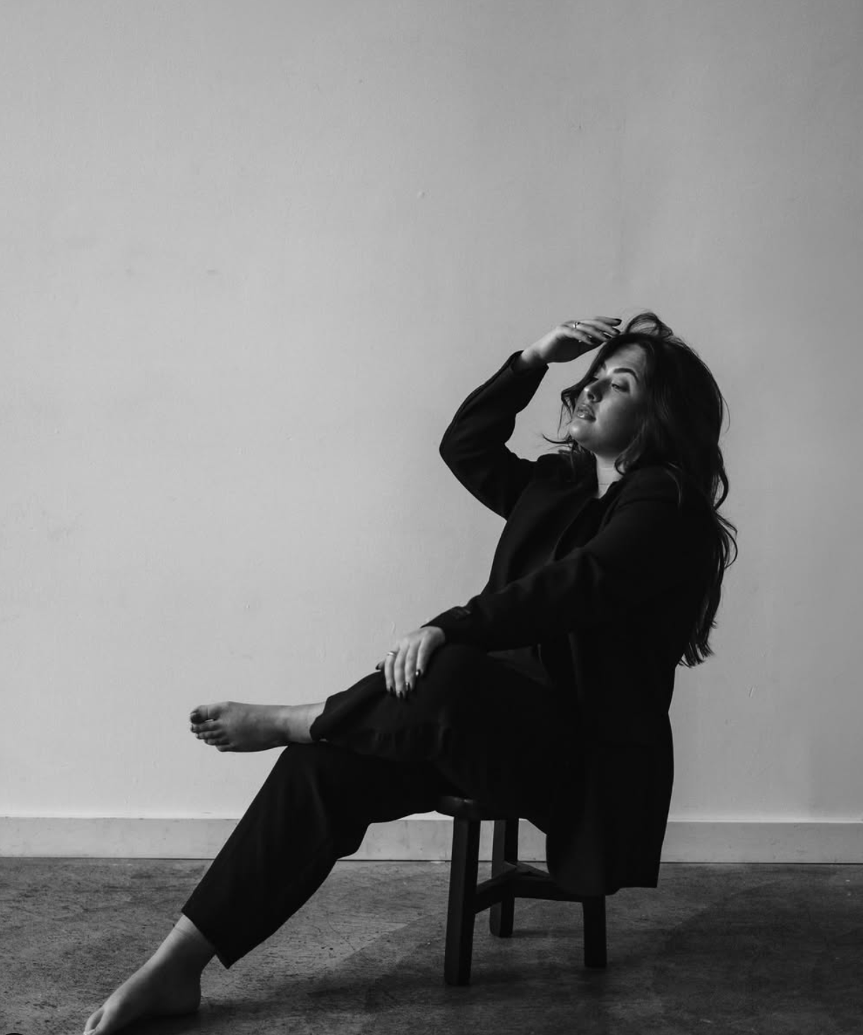 Dramatic black and white portrait of person in dark clothing seated on chair with hand raised.