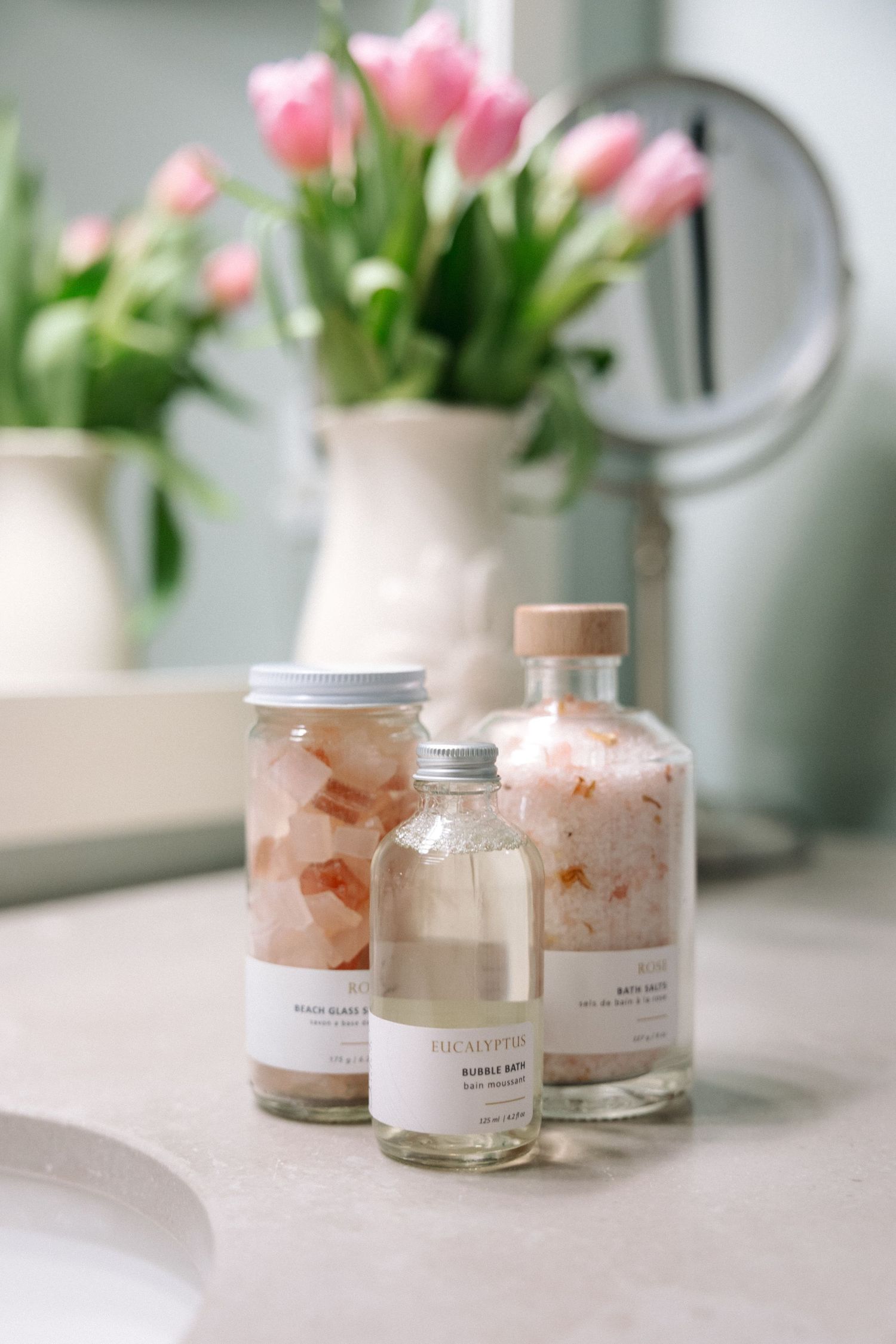 Three bath product bottles with pink bath salts on a marble countertop, with pink tulips in a white vase behind.