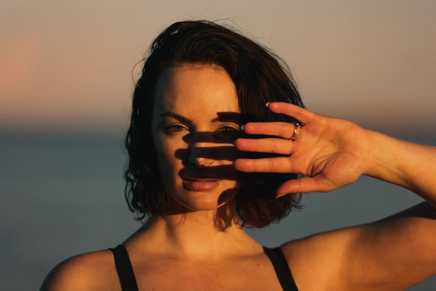 Artistic portrait of a person shielding their face with their hand against a warm sunset backdrop.