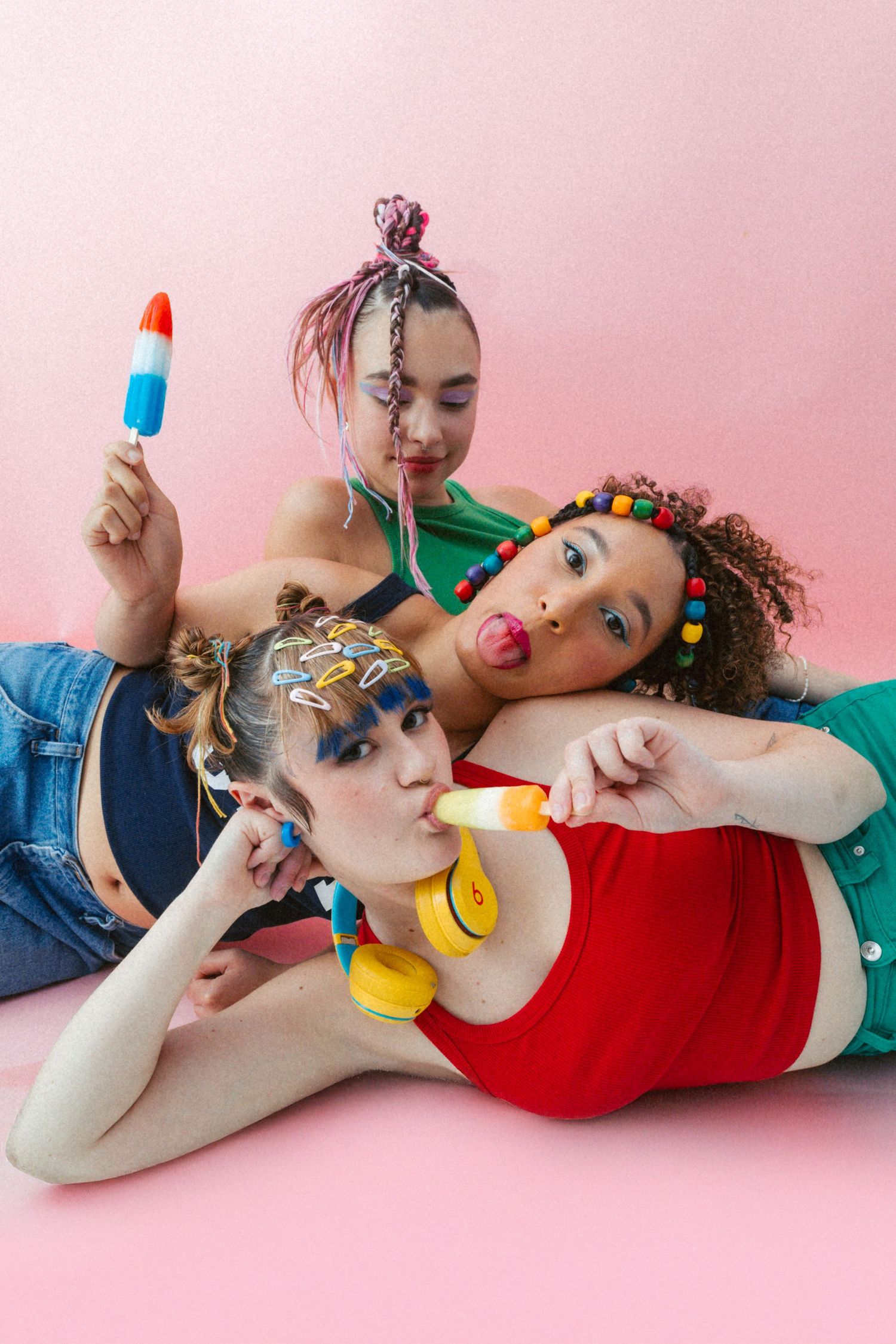 Three friends pose playfully with popsicles and colorful accessories against a pink backdrop.