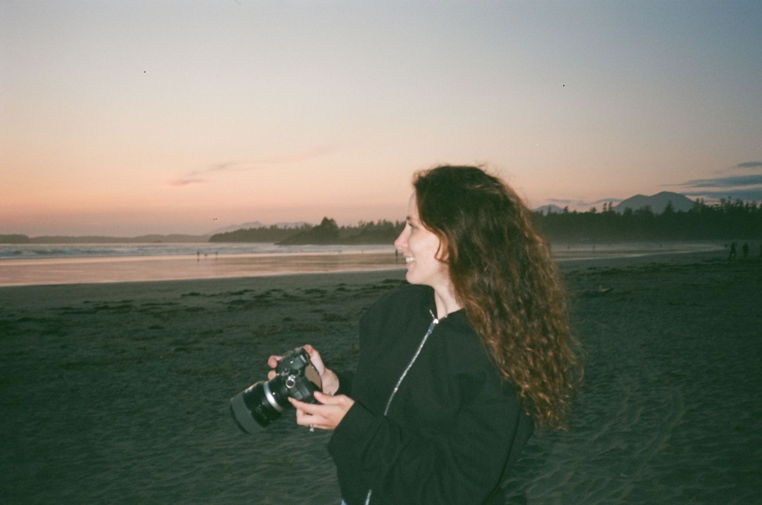 A person with long wavy hair holds a camera on a beach at sunset.