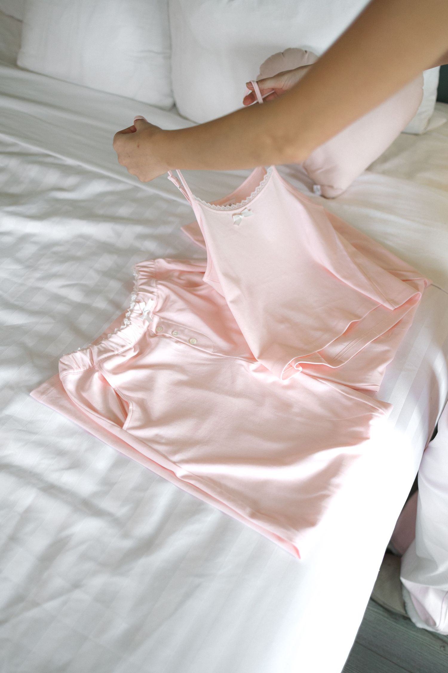 Pink satin pajama set with lace trim laid on white bed, with woman sitting nearby holding matching pink item.