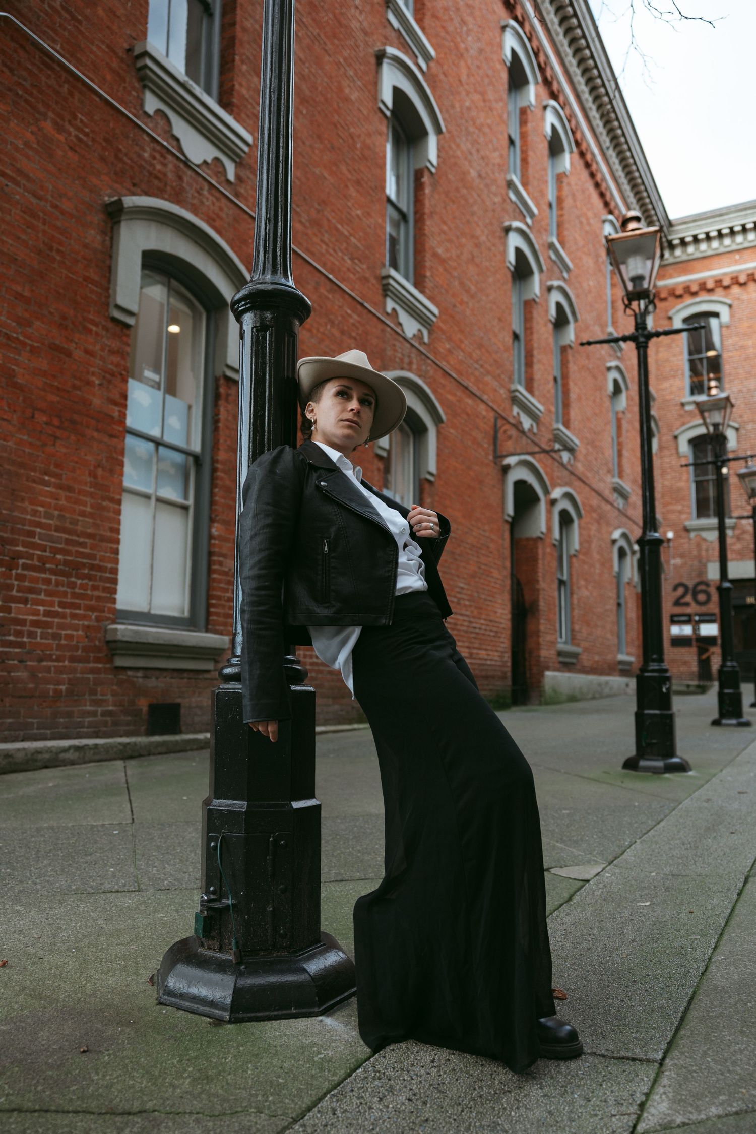Two people in formal black attire pose dramatically against a brick building with arched windows and lamp posts.