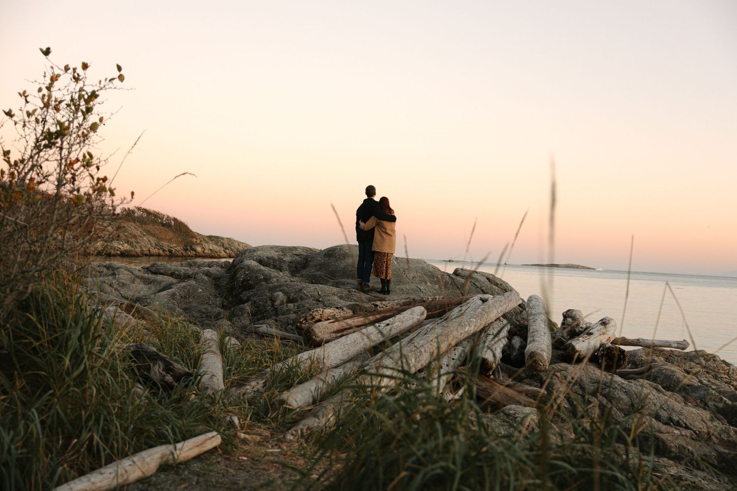 A silhouetted couple stands on driftwood-covered rocks at sunset with a sailboat visible in the distance.