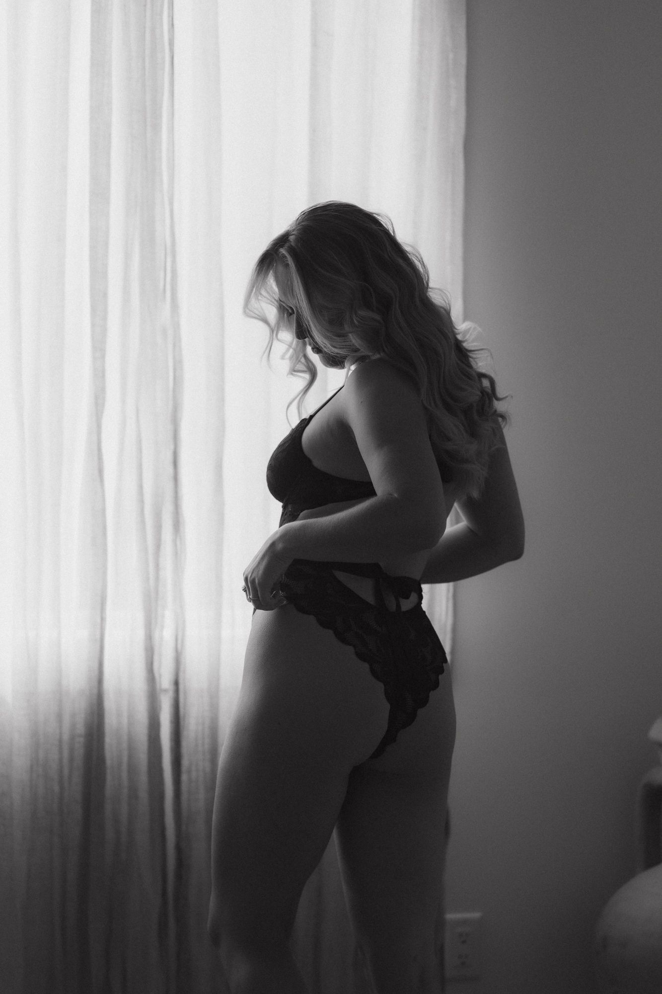 Black and white artistic boudoir silhouette of a woman in lingerie standing by a window with soft lighting.
