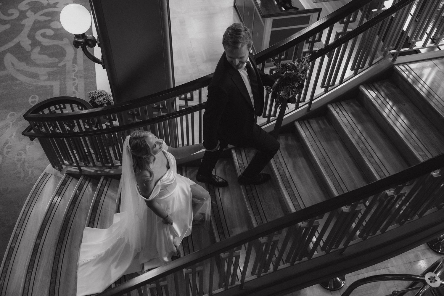 A black and white photograph of a couple ascending an elegant curved staircase with ornate railings.