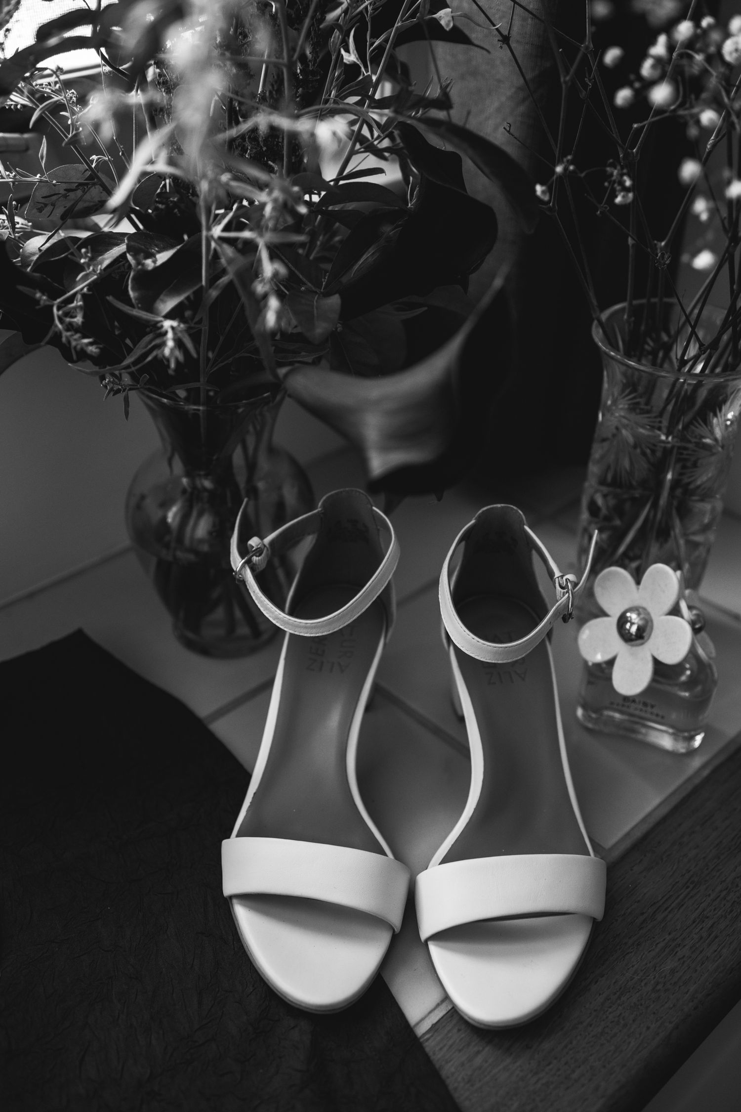 Elegant white bridal sandals displayed with delicate flowers in vases on a dark wooden surface.
