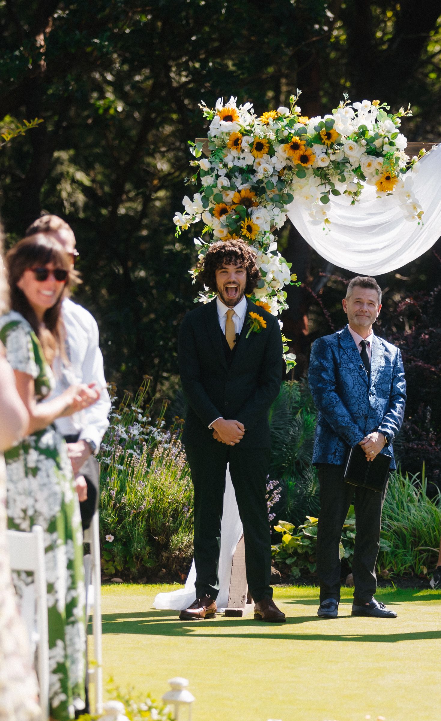 Outdoor wedding ceremony with floral arch and guests gathered in garden setting.