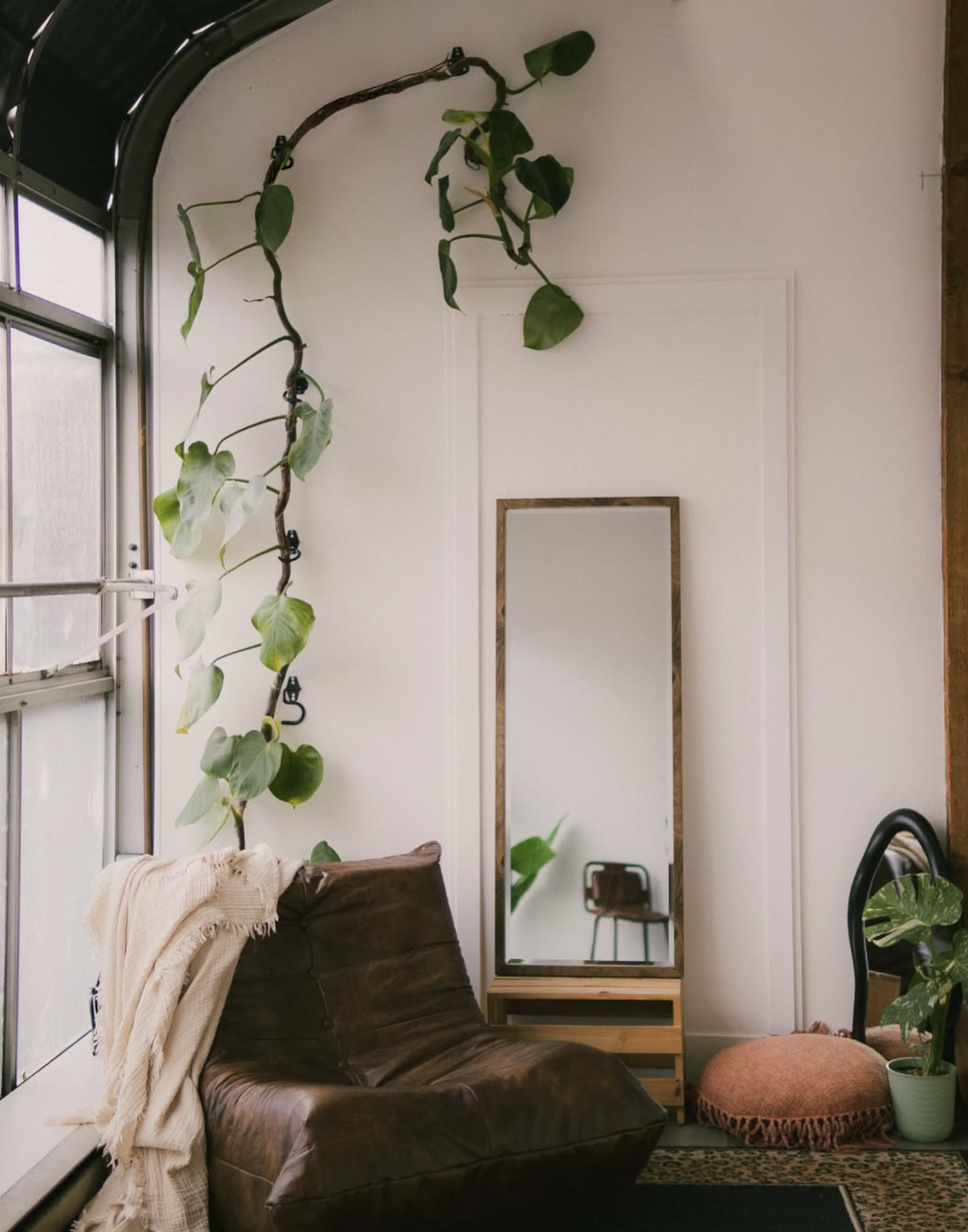 Cozy living room corner with a climbing vine plant, brown leather sofa, floor mirror, and boho pouf on a patterned rug.