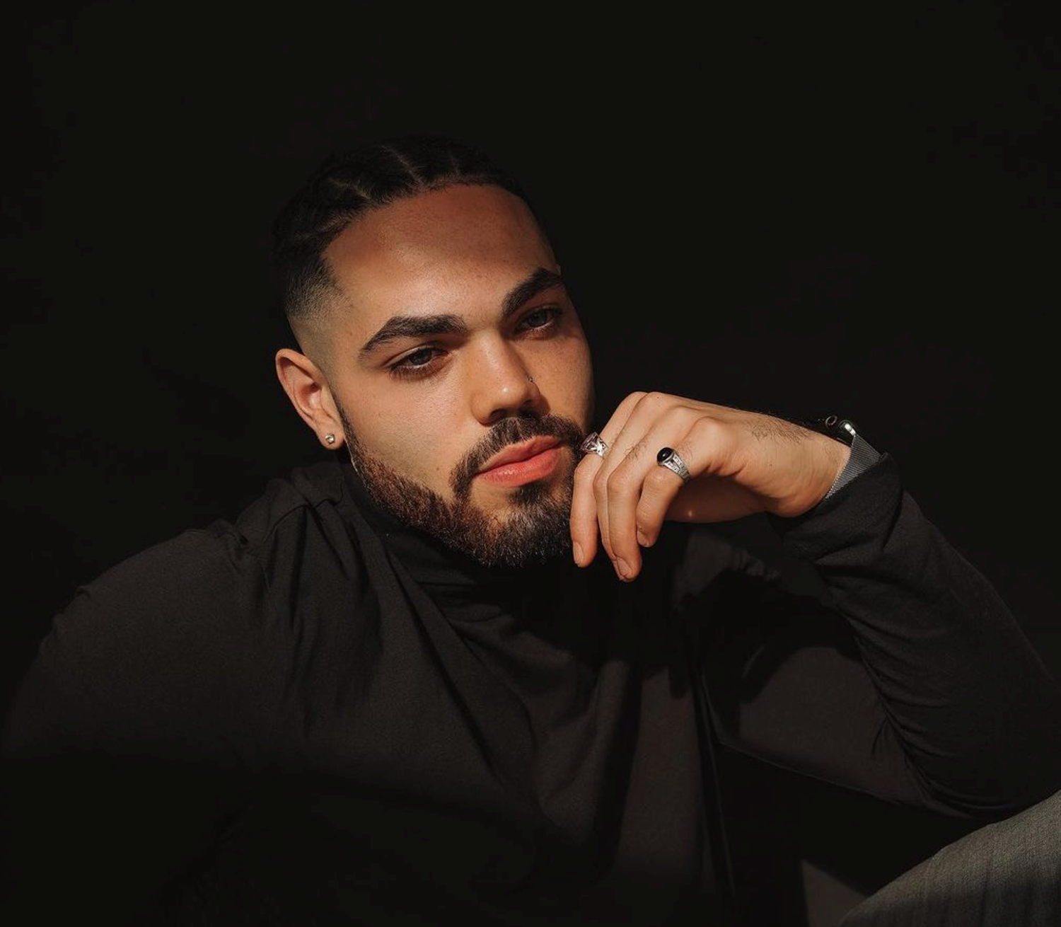 Studio portrait of a person in a black shirt with facial hair posing against a dark background, hand thoughtfully near face.