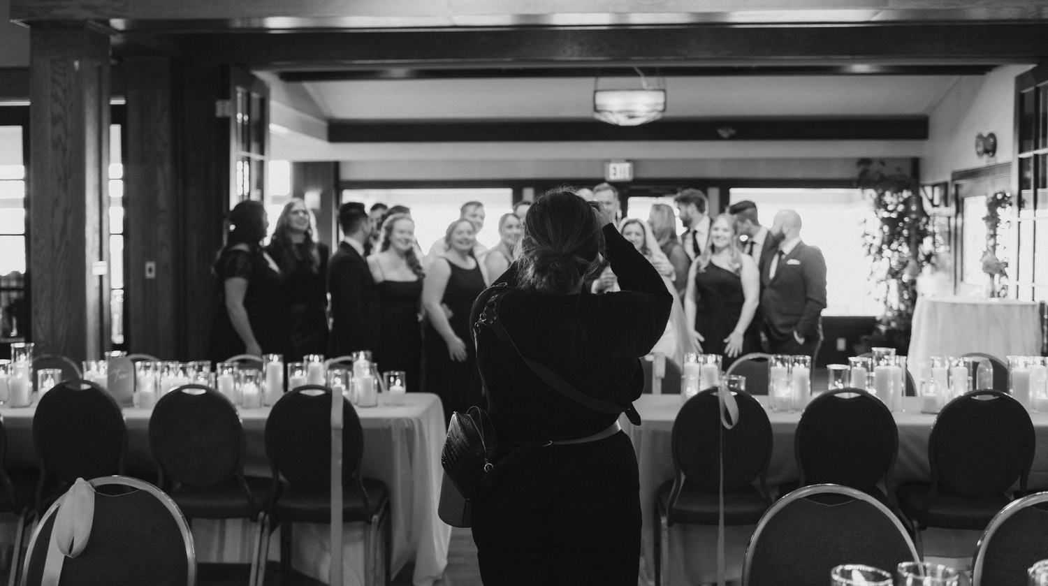 A black and white photo of a banquet hall with tables and people standing in the background.