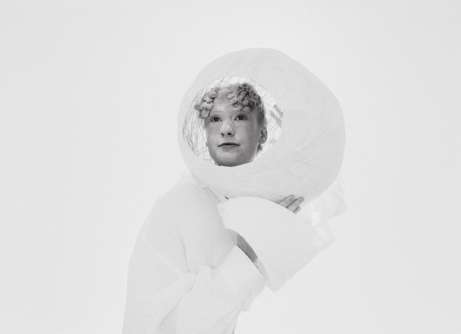 Black and white photo of a person peering through a circular white sculptural piece, wearing a white outfit against a white background.