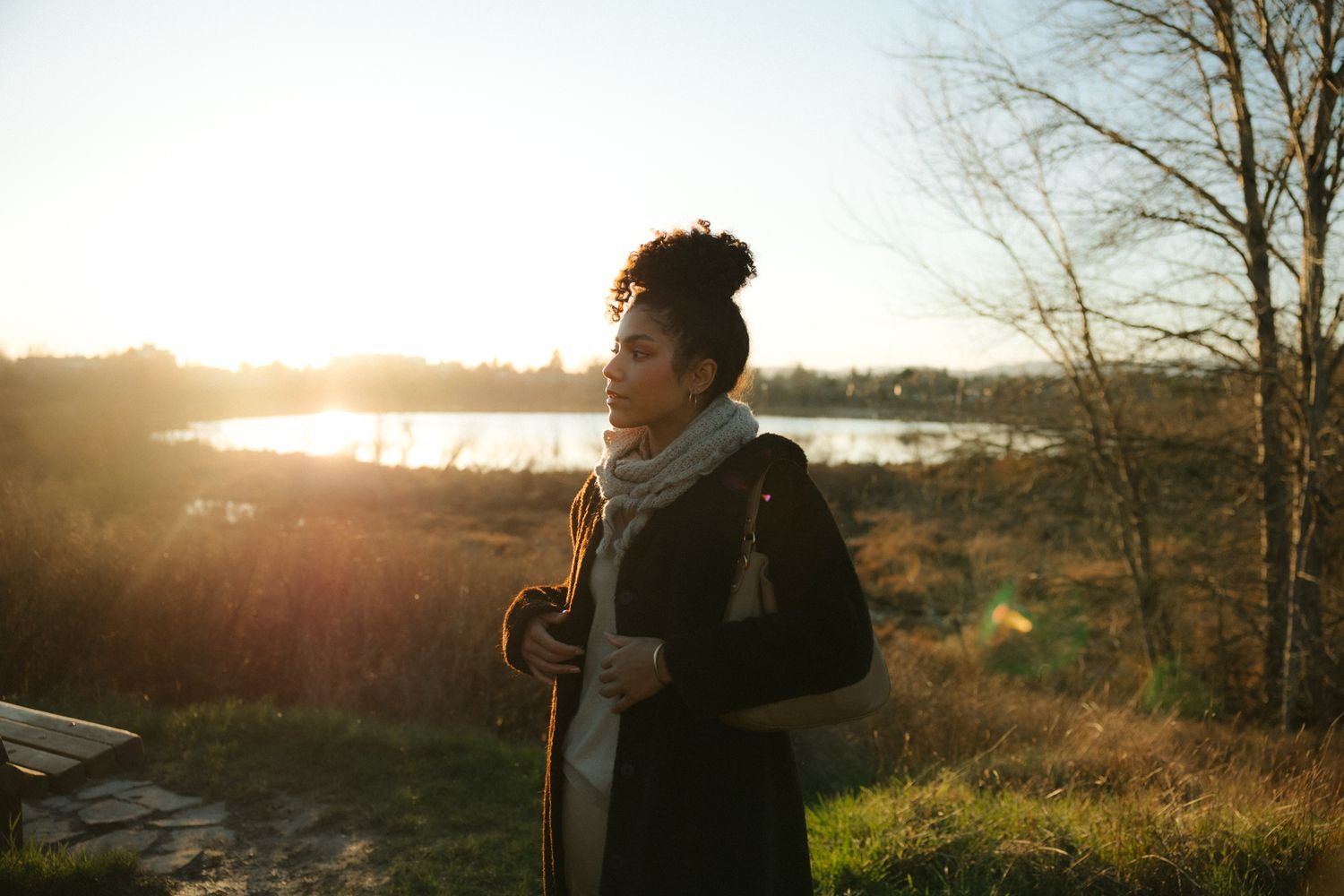 Woman in coat and scarf standing outdoors at golden hour near a lake surrounded by autumn trees.