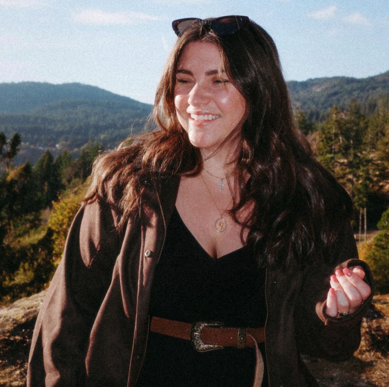 Smiling young woman with long dark hair wearing a brown jacket and belt, posing outdoors with scenic mountain views.
