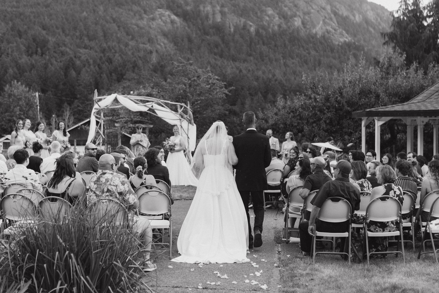 Black and white outdoor wedding ceremony with bride and groom walking down the aisle toward a floral arch.