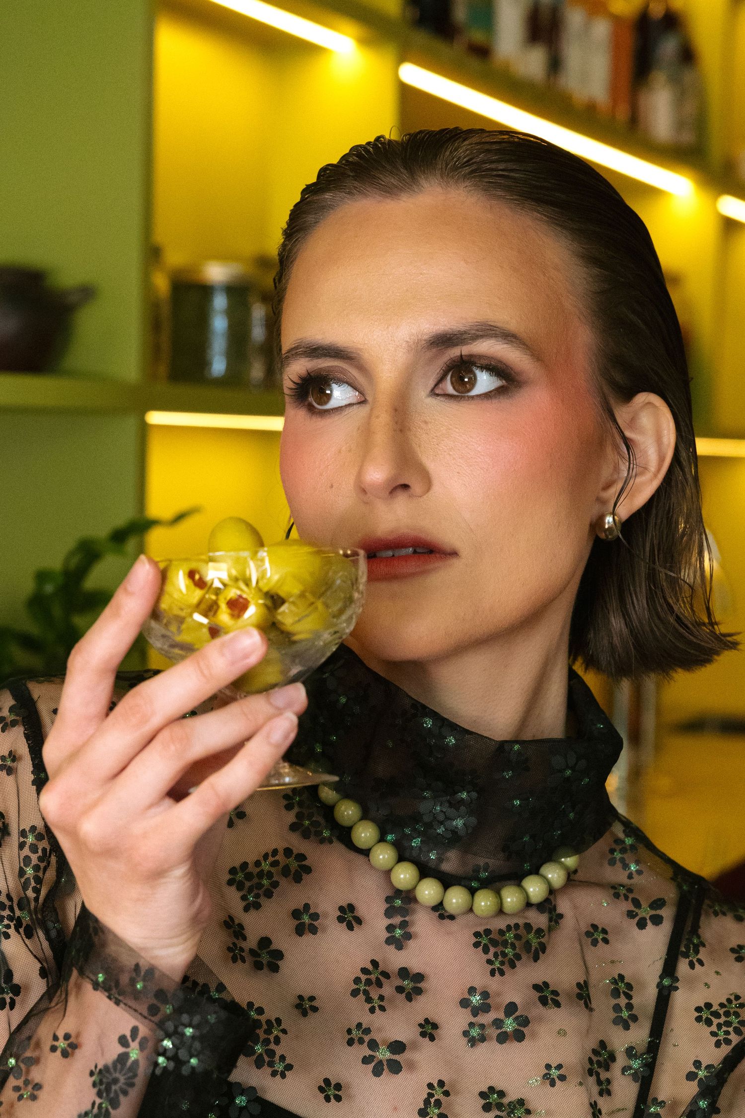 Woman in sheer floral top sipping a green olive martini cocktail in a stylish bar with yellow lighting.