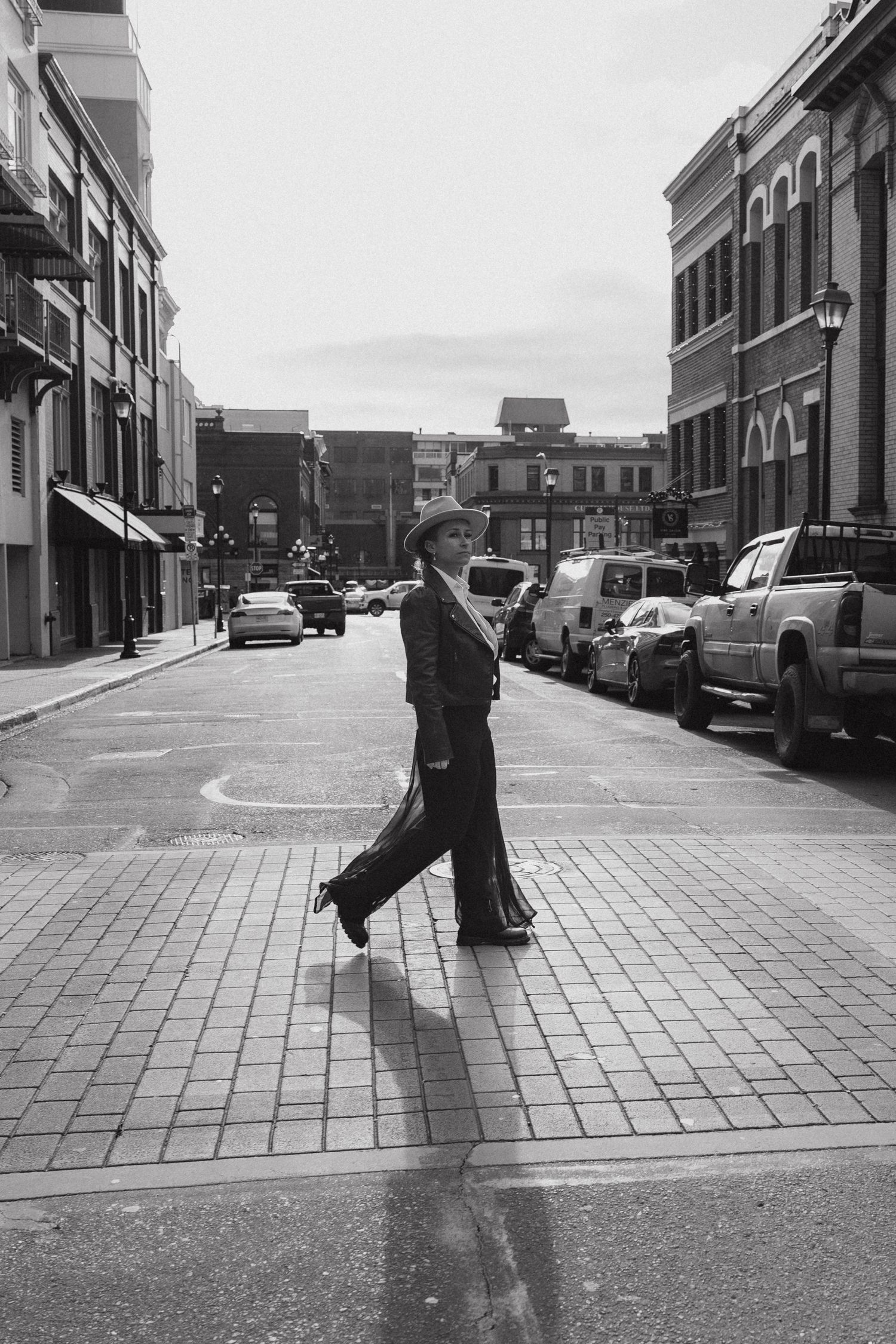 A person walks across a brick-paved street in a black and white urban scene with parked cars and historic buildings.