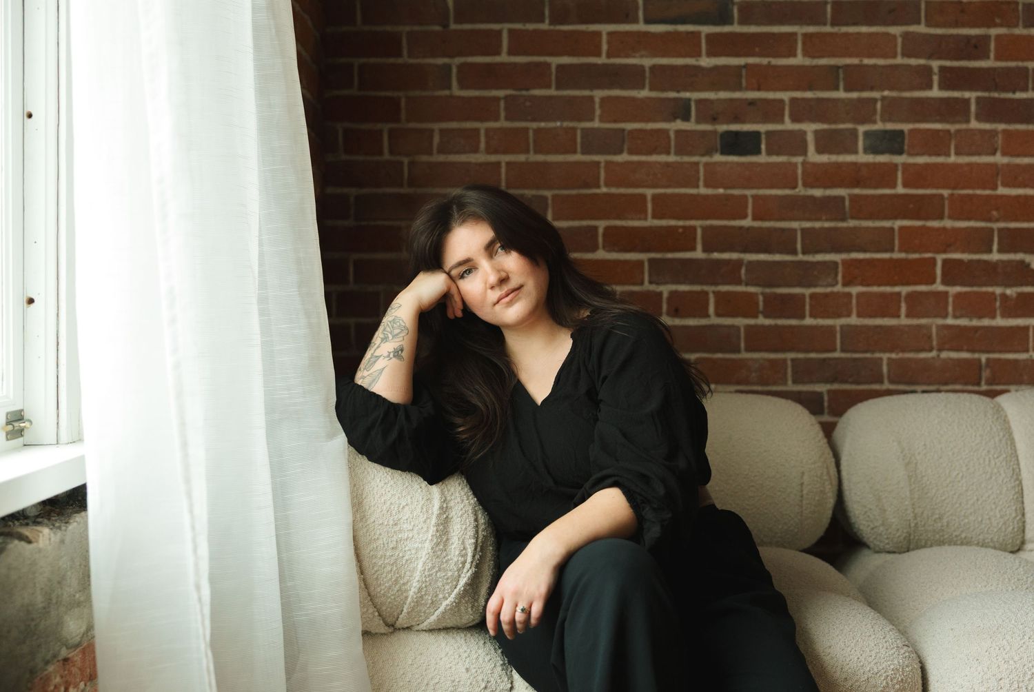 A person in a black sweater poses casually on a gray couch against a brick wall in a bright room.