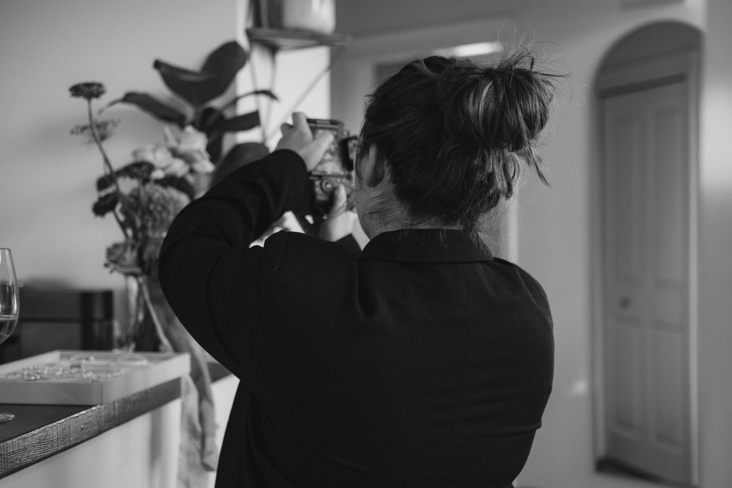 A black and white photo shows a person with messy hair photographing a houseplant in an interior setting.