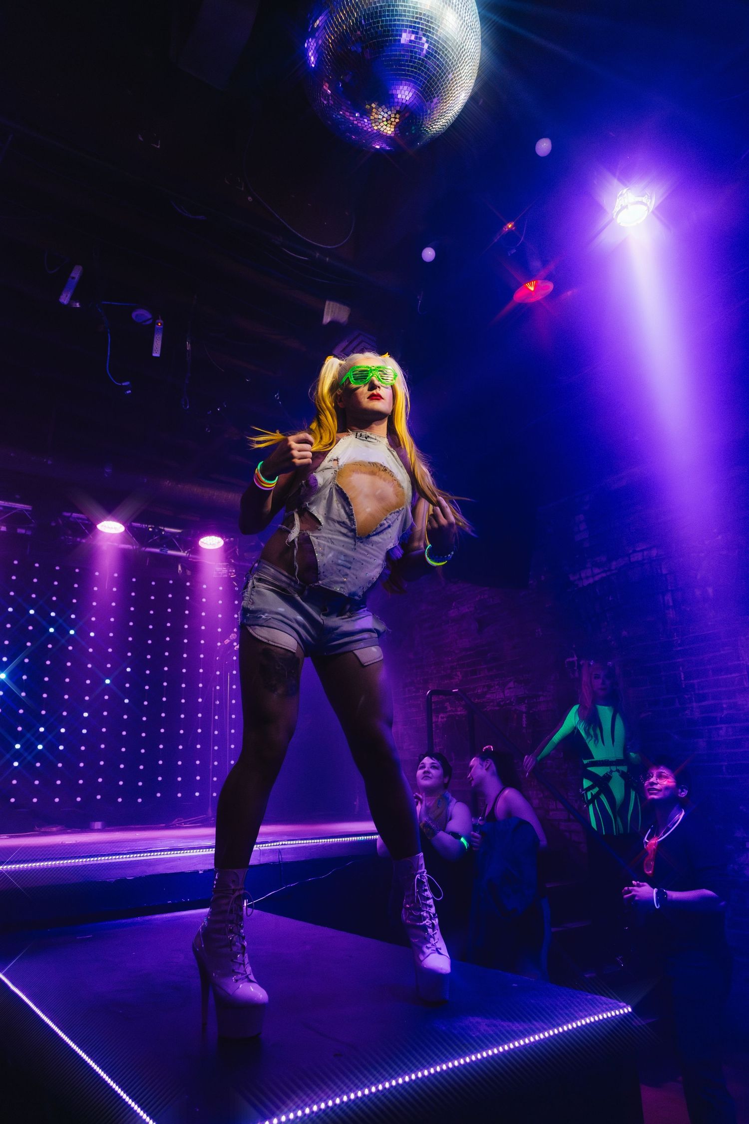 Performer in neon lighting dances on stage beneath disco ball at nightclub.