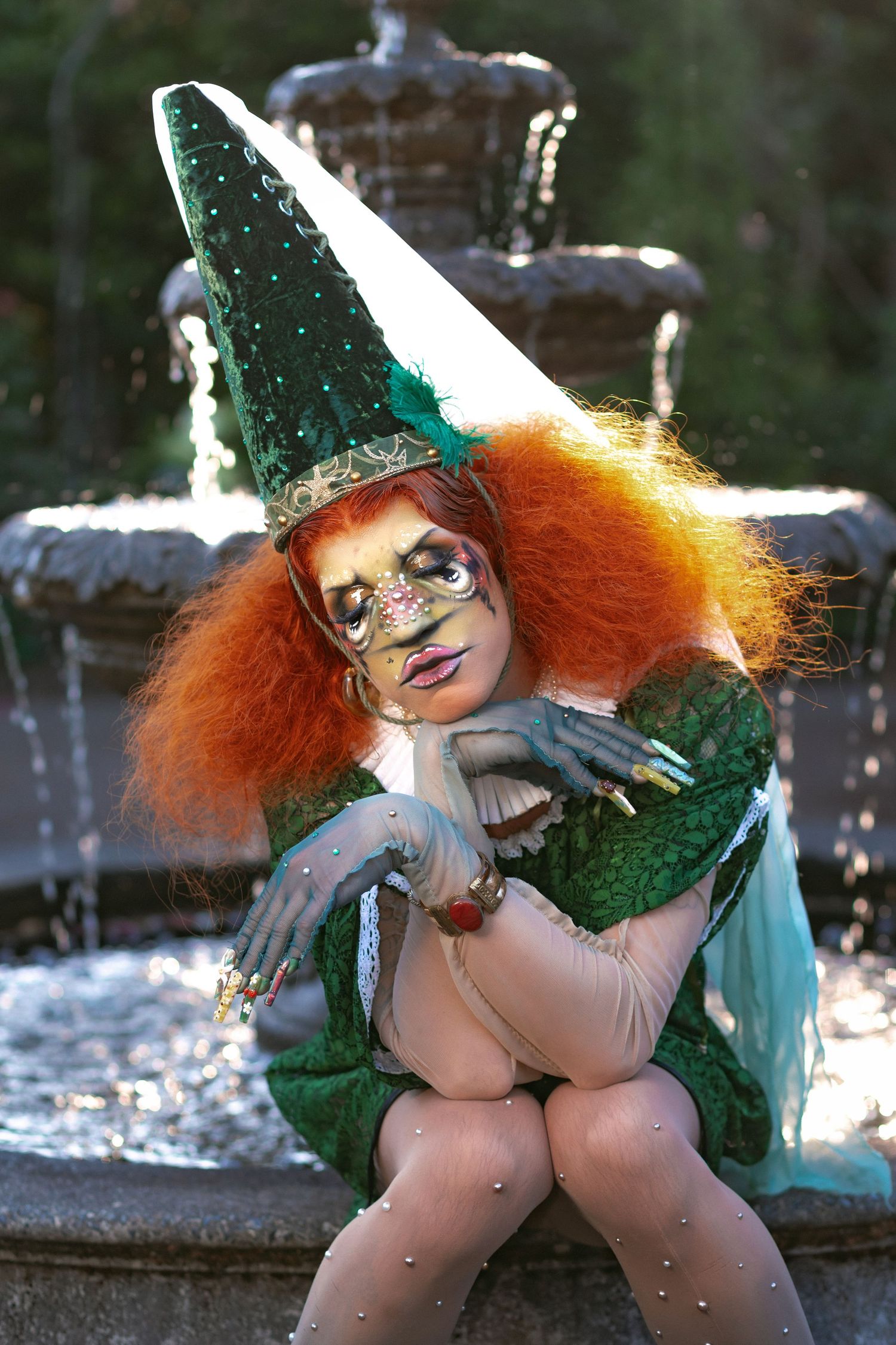 Whimsical fantasy character in sparkly green witch costume with bright orange hair poses playfully by fountain.