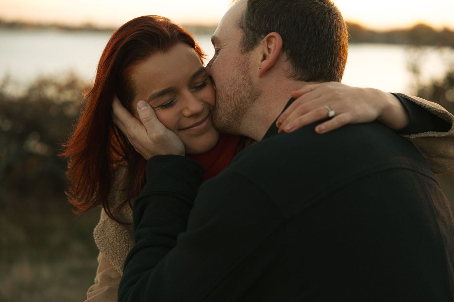 A man tenderly kisses a red-haired woman on the cheek at sunset near a waterfront, as she smiles warmly.