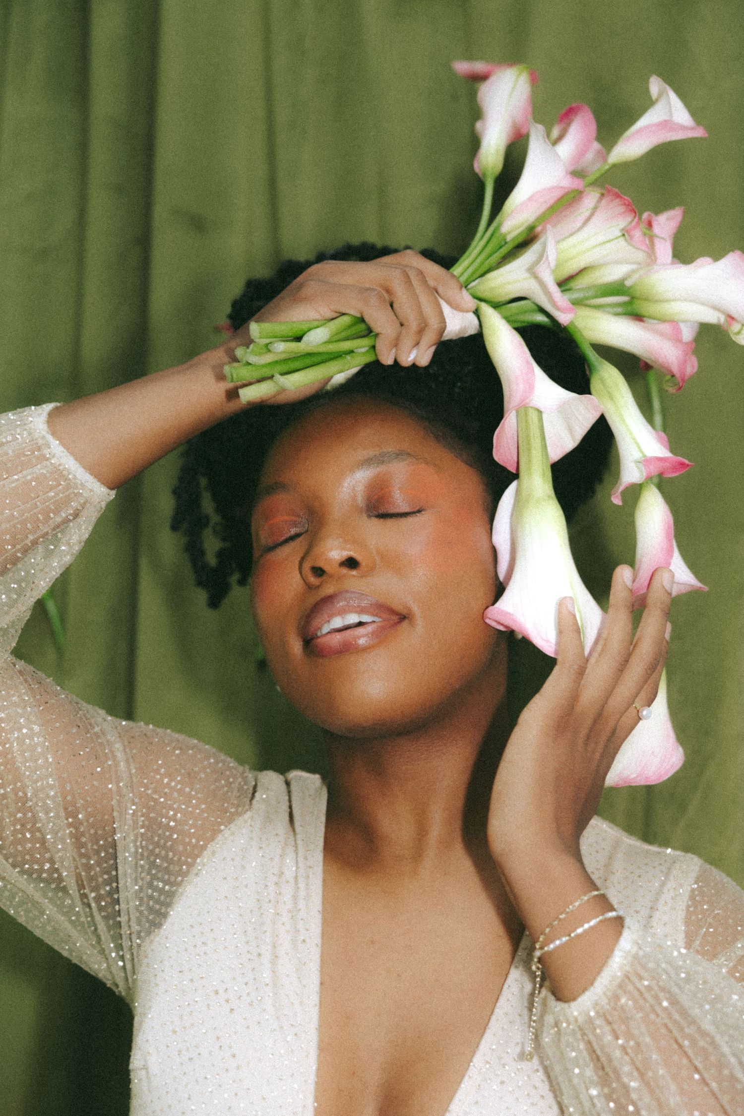 A Black woman in a white sequined dress holds pink calla lilies above her head with eyes closed against a green backdrop.
